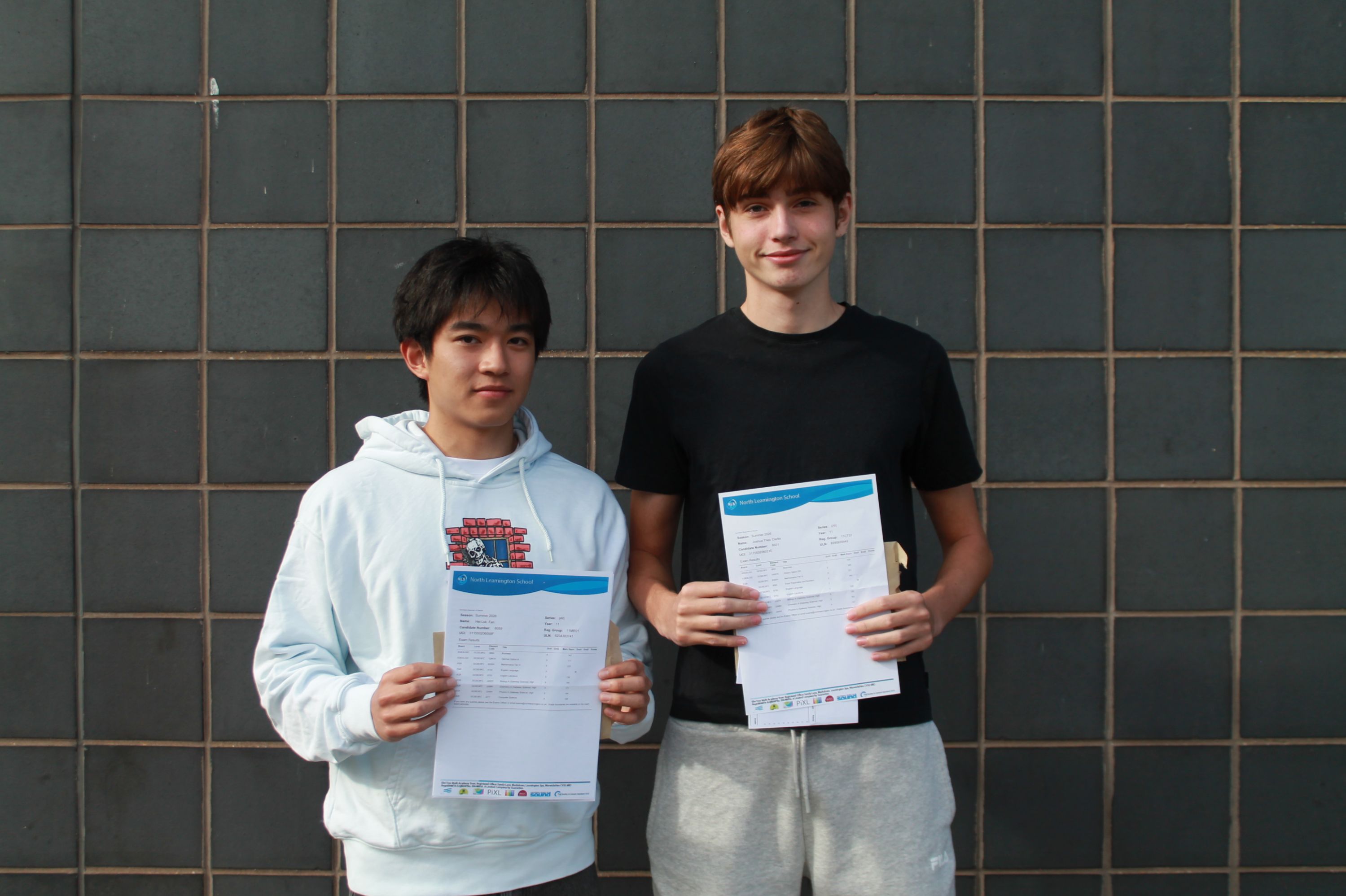 Photograph of two young men standing side by side against a tiled wall, each holding documents or envelopes