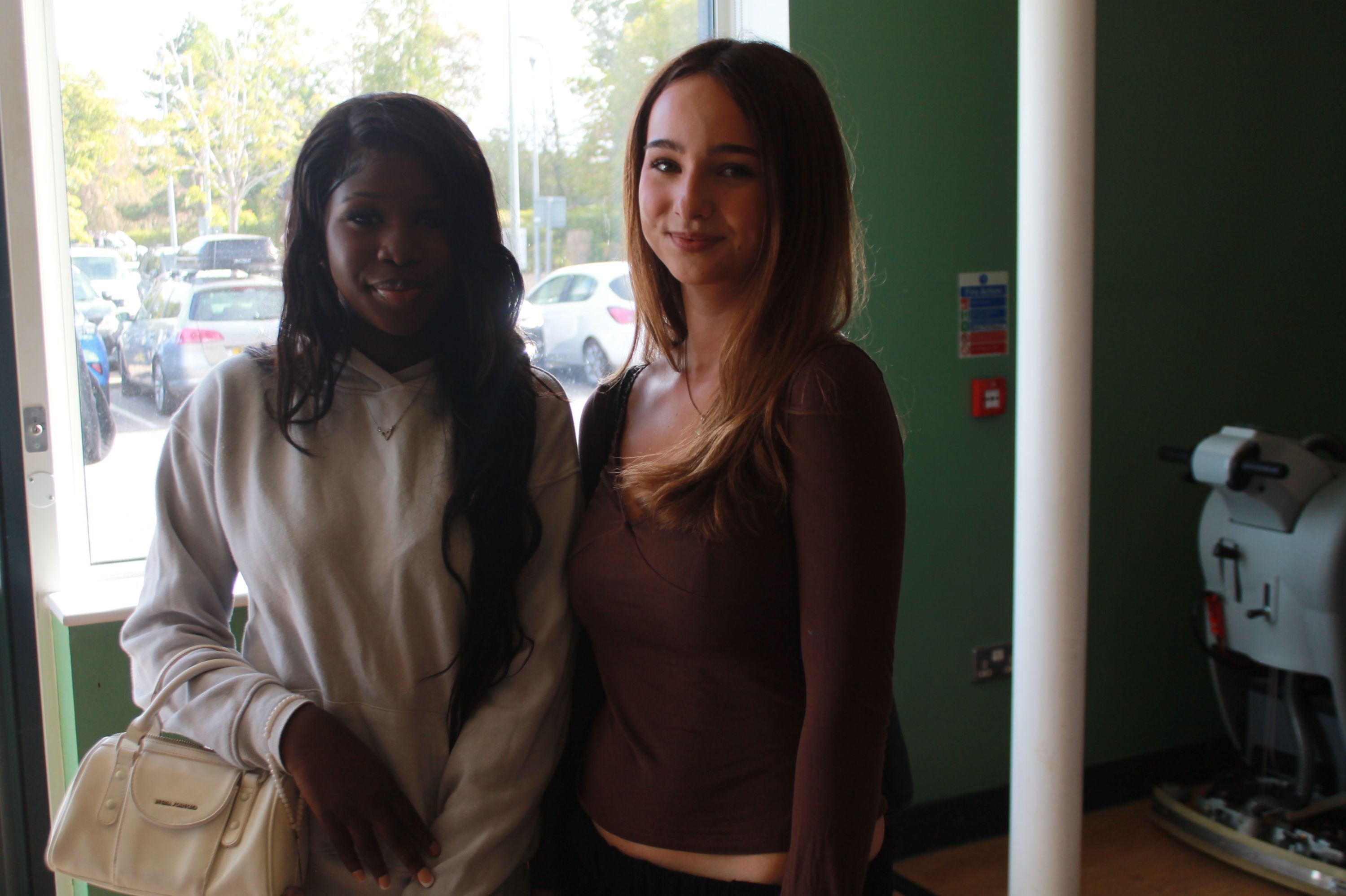 Photograph of two young women standing indoors near a window with parked cars visible outside. One woman wears a white hoodie and holds a white handbag, while the other wears a brown long-sleeve top, both smiling at the camera.