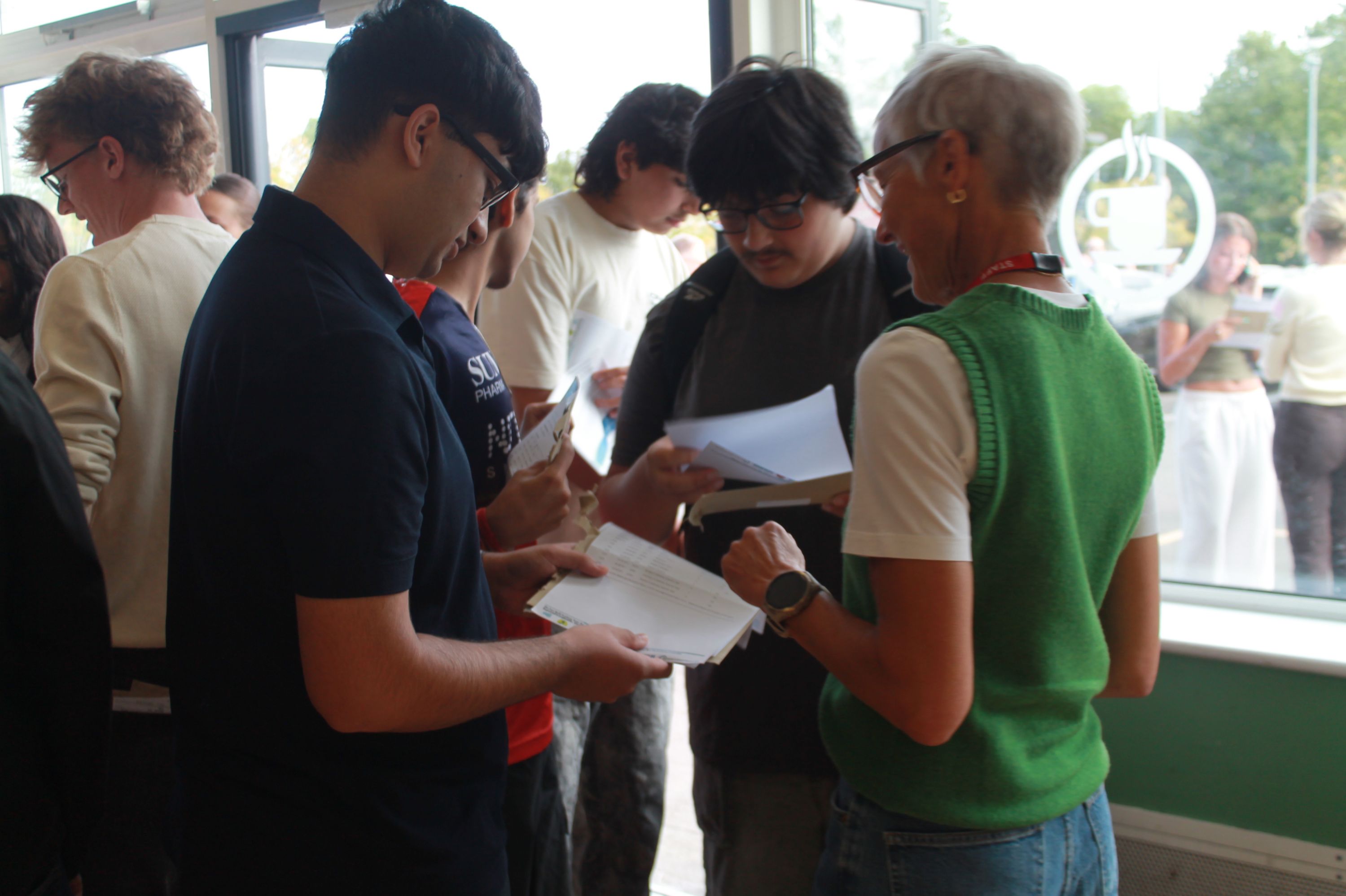 ​​Photograph showing a group of people indoors engaged in conversation and reviewing papers, 
