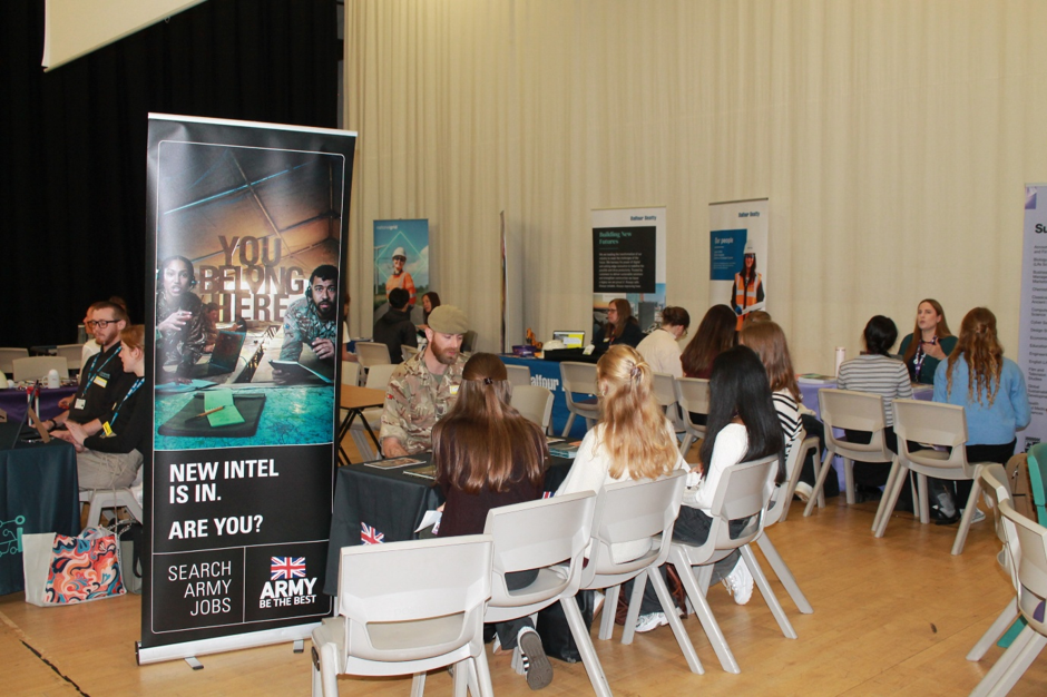 Photograph of an Army recruitment and information event, showing attendees speaking with uniformed personnel at tables inside a hall. Foreground banner reads “YOU BELONG HERE” and “NEW INTEL IS IN. ARE YOU?”, with British Army branding, Union Jack, presentation screens, brochures and displays