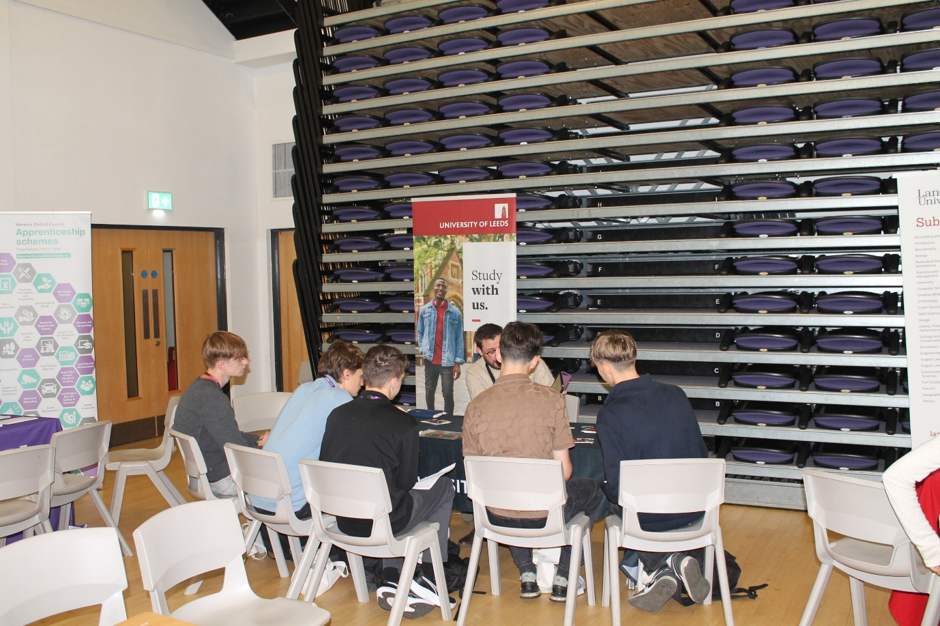 Photograph shows five  students seated in a group study or discussion area beside rows of purple tiered seats. University of Leeds promotional banner, event posters, chairs and illuminated exit sign 