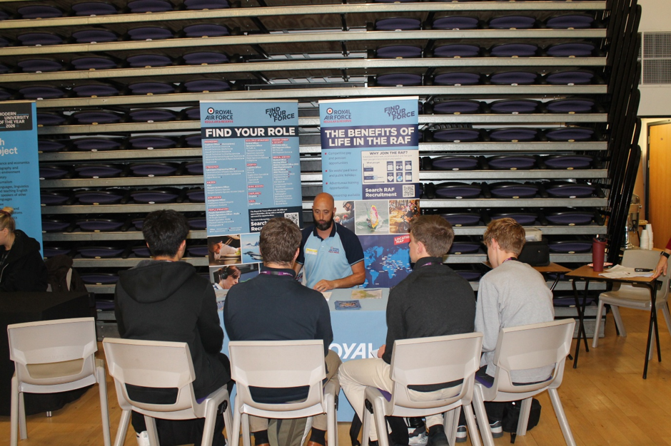 Photograph of a Royal Air Force careers presentation, with seated young attendees listening to a uniformed recruiter. Blue and white display banners read “Find Your Role” and “The Benefits of Life in the RAF”, with career information panels and RAF branding.