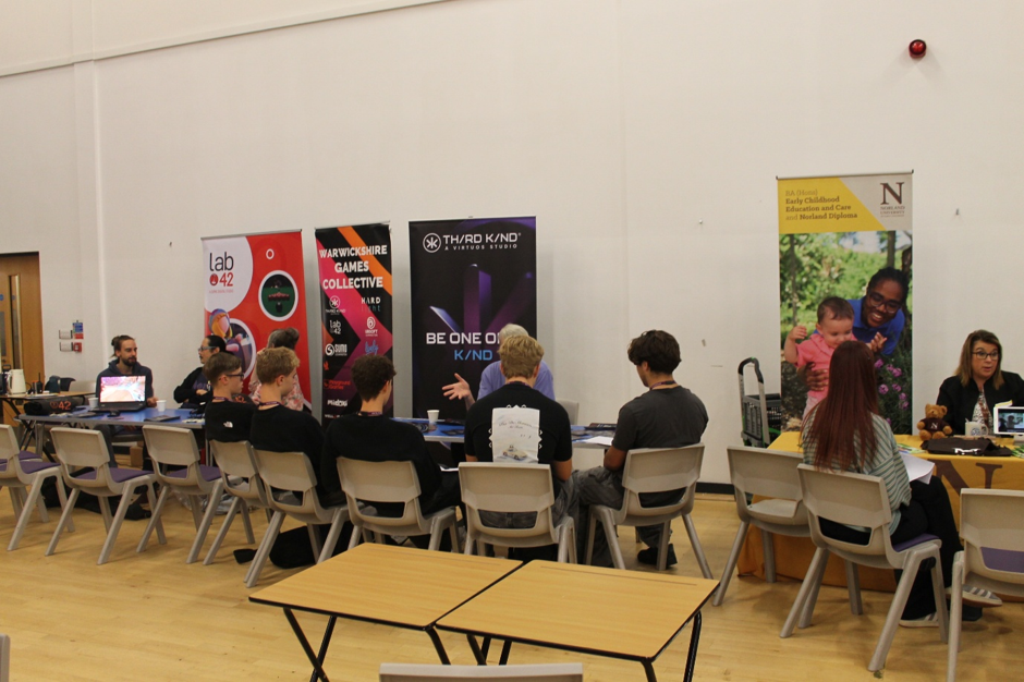 Photograph showing a careers event, with students seated at tables speaking with exhibitors. Visible banners promote Lab 842, Games Collective, Third Kind Games and creative opportunities, while additional staff and information stands line rear wall.