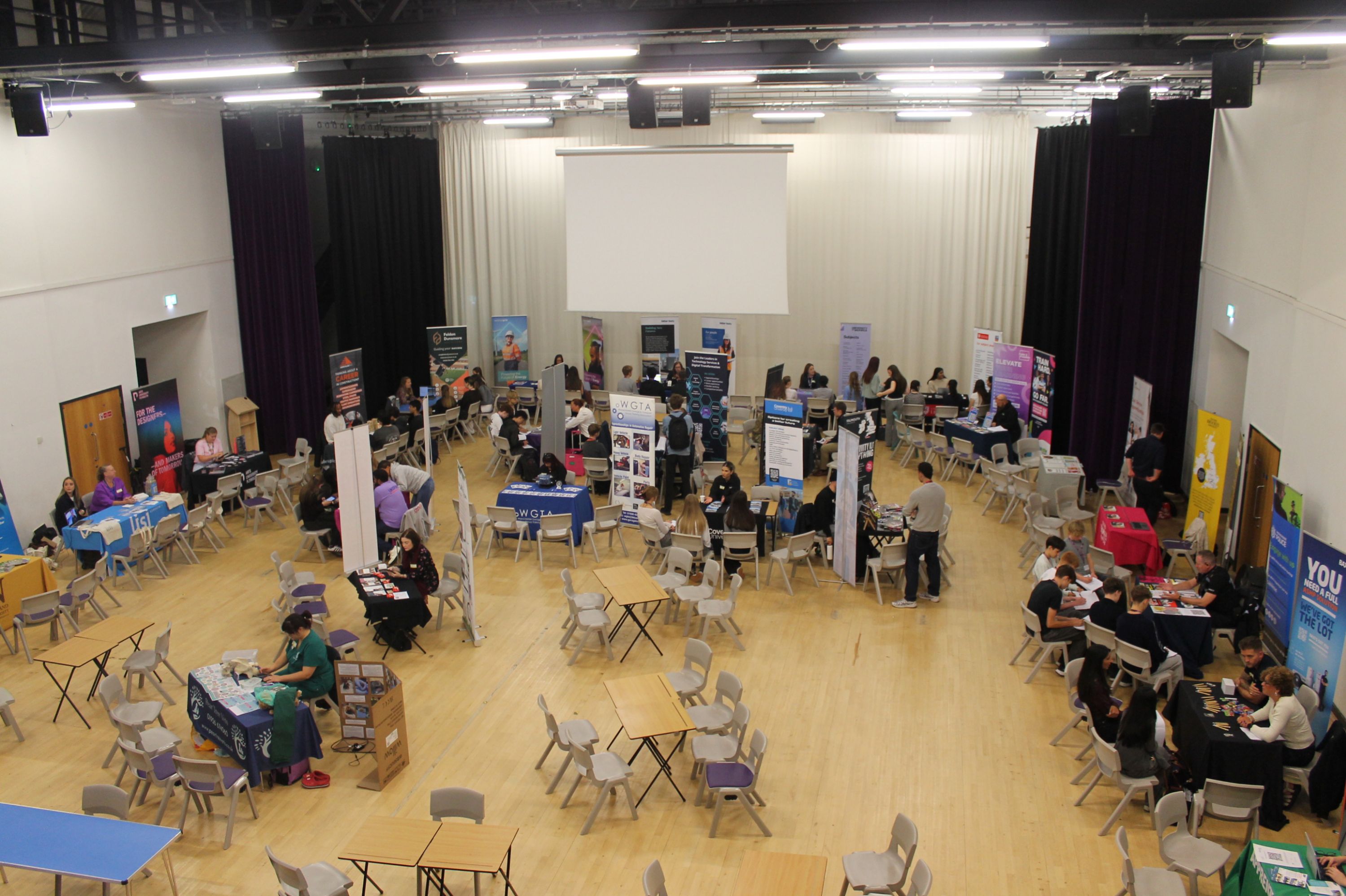 Photograph shows a busy careers fair inside a large hall, with numerous exhibitors, information stands and seated visitors. Open floor layout, timber flooring, black curtains, projection screen, colourful banners and clusters of attendees highlight networking, advice and recruitment activity.