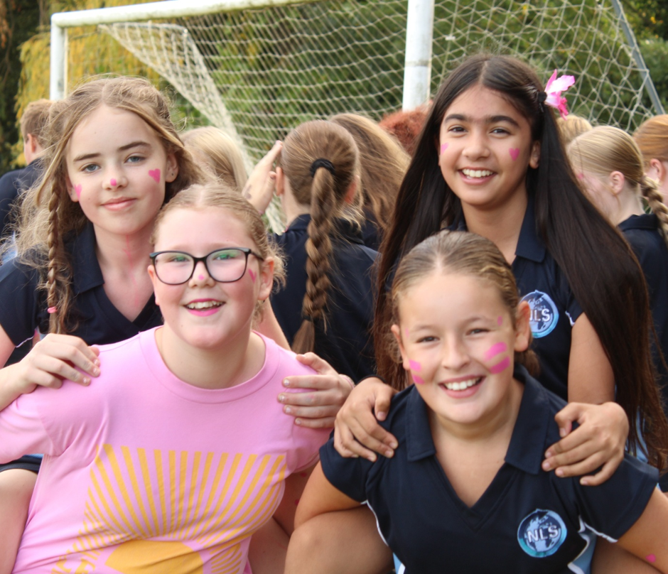 Photograph showing four smiling schoolgirls posing together beside a football goal, likely during Race for Life Event event. Girls wear navy uniforms and pink face paint, while one wears glasses and a pink top; football net and other pupils form background.