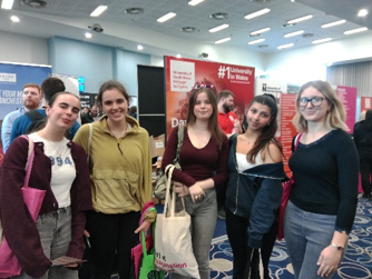 Photograph of five young women posing at a university careers  event. University in Wales and promotional banners form backdrop, with branded tote bags, clothing and exhibition stands indicating an educational outreach setting.