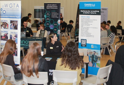 Photograph shows a busy careers and fair, with students and exhibitors meeting around tables. Branded information stands, including WGTA and Community IT, display technology, training and sustainability opportunities, while attendees browse leaflets and talk with representatives.