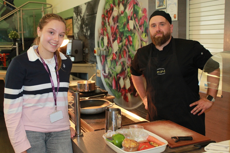 Photograph showing two people posing beside a counter in a café . Stainless-steel pans, serving equipment, fresh ingredients and plated food appear in foreground, with colourful mural and kitchen fittings behind.
