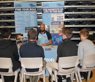Photograph shows an RAF careers information session, with a uniformed representative speaking to four seated attendees. Promotional boards labelled “Find Your Role” and “Benefits of Life in RAF” display recruitment information beside rows of empty seats and aviation-themed visuals.
