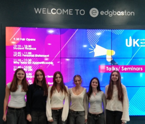 Photograph of six young women posing in front of a colourful Edgbaston welcome display for a UK student event. Digital screen shows talks and seminars, a timetable, UK branding, bright blue and magenta graphics, and event welcome text.