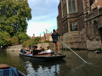 Photograph showing a guided punting tour along a narrow canal beside historic brick buildings. A standing guide steers a flat-bottomed boat carrying seated passengers, with trees, reflections and additional boats visible.