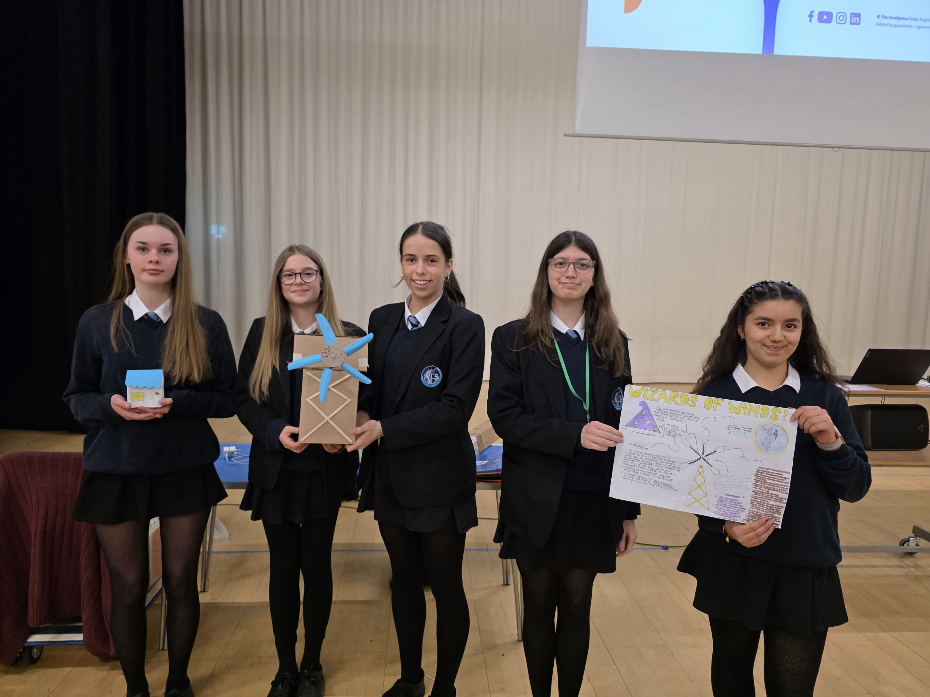 Photograph of five students in uniform  holding a small model, a wind turbine project, and a poster with drawings and tex