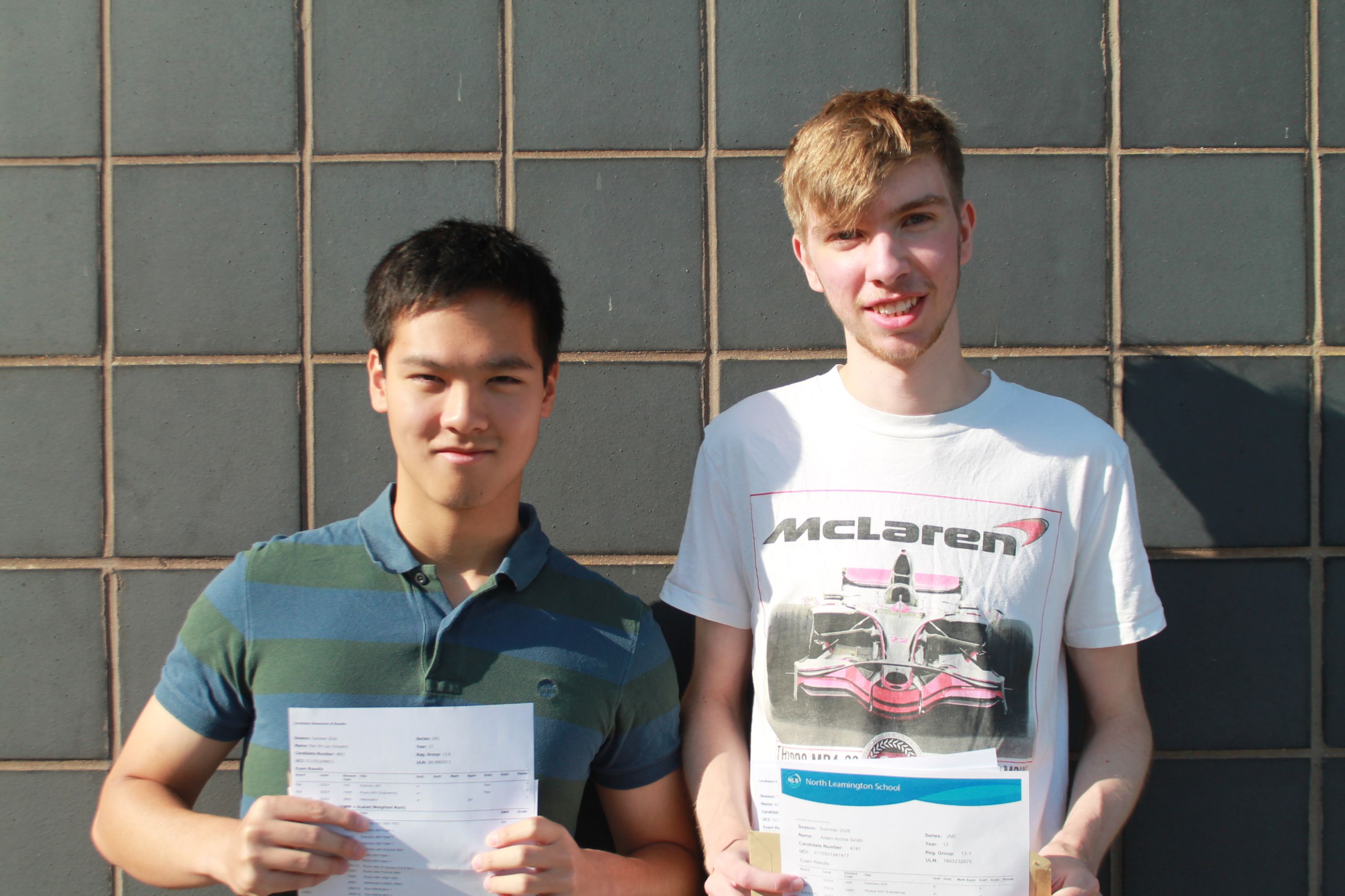 Photograph of two young men standing side by side against a tiled wall, each holding a document with their A Level results with one wearing a striped polo shirt and the other a white McLaren-themed t-shirt.