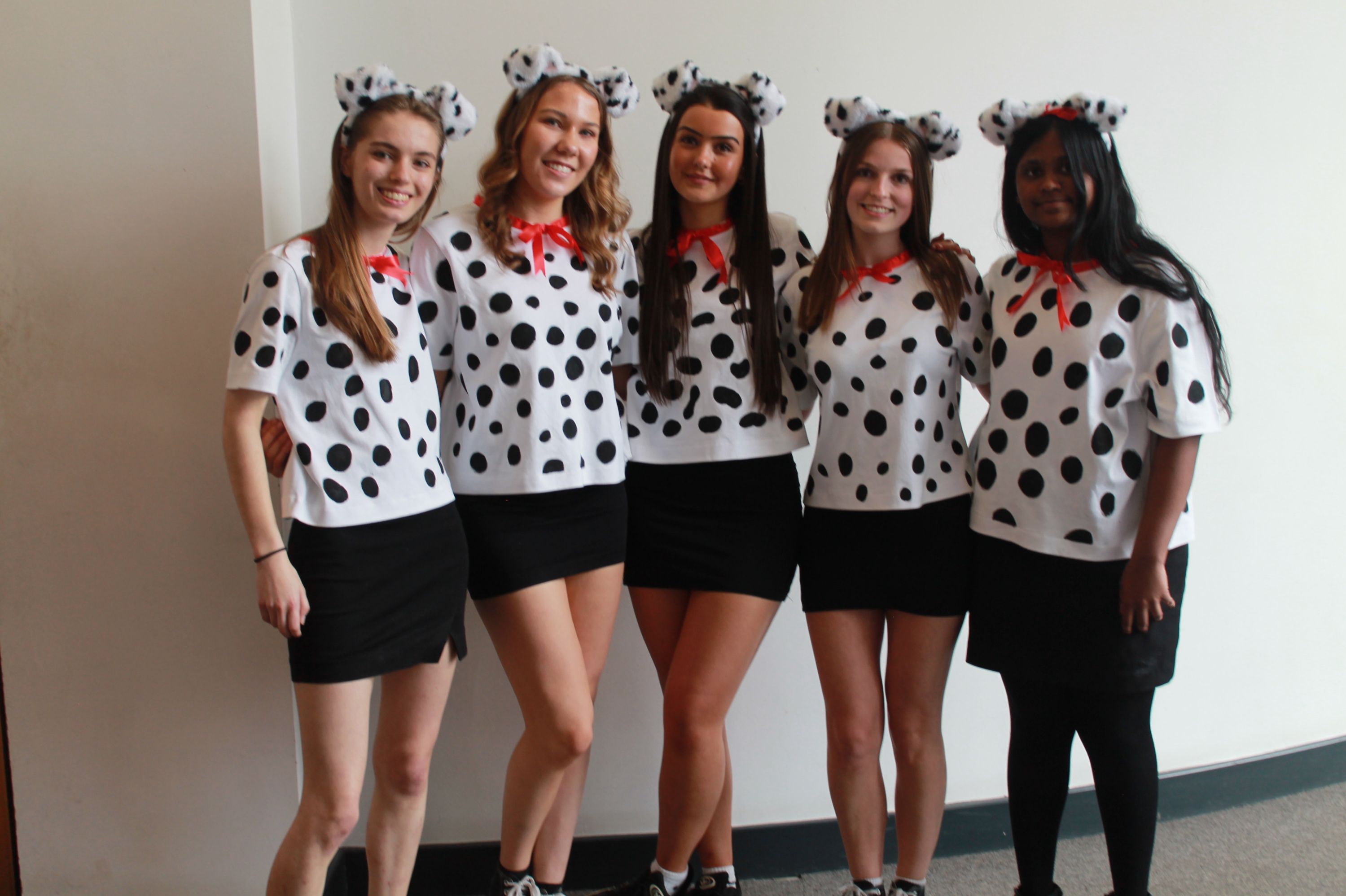 Photograph of five people dressed in matching Dalmatian-themed costumes, featuring white tops with black spots, black skirts, red neck ribbons, and Dalmatian ear headbands