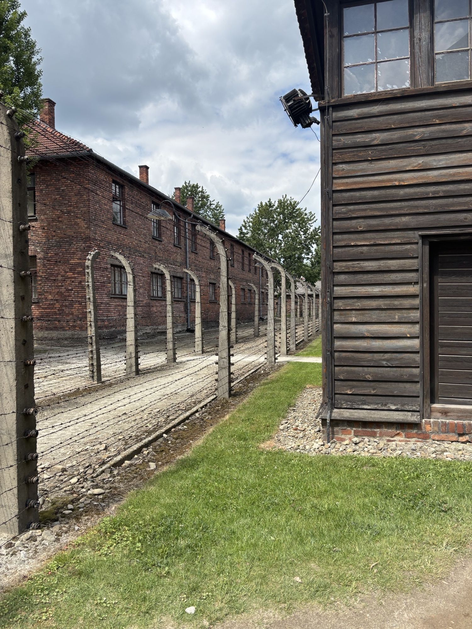 Photograph showing a section of a historical concentration camp with barbed wire fences and brick buildings. The scene highlights security structures including multiple rows of fencing and watchtowers, set against a partly cloudy sky and grassy area.