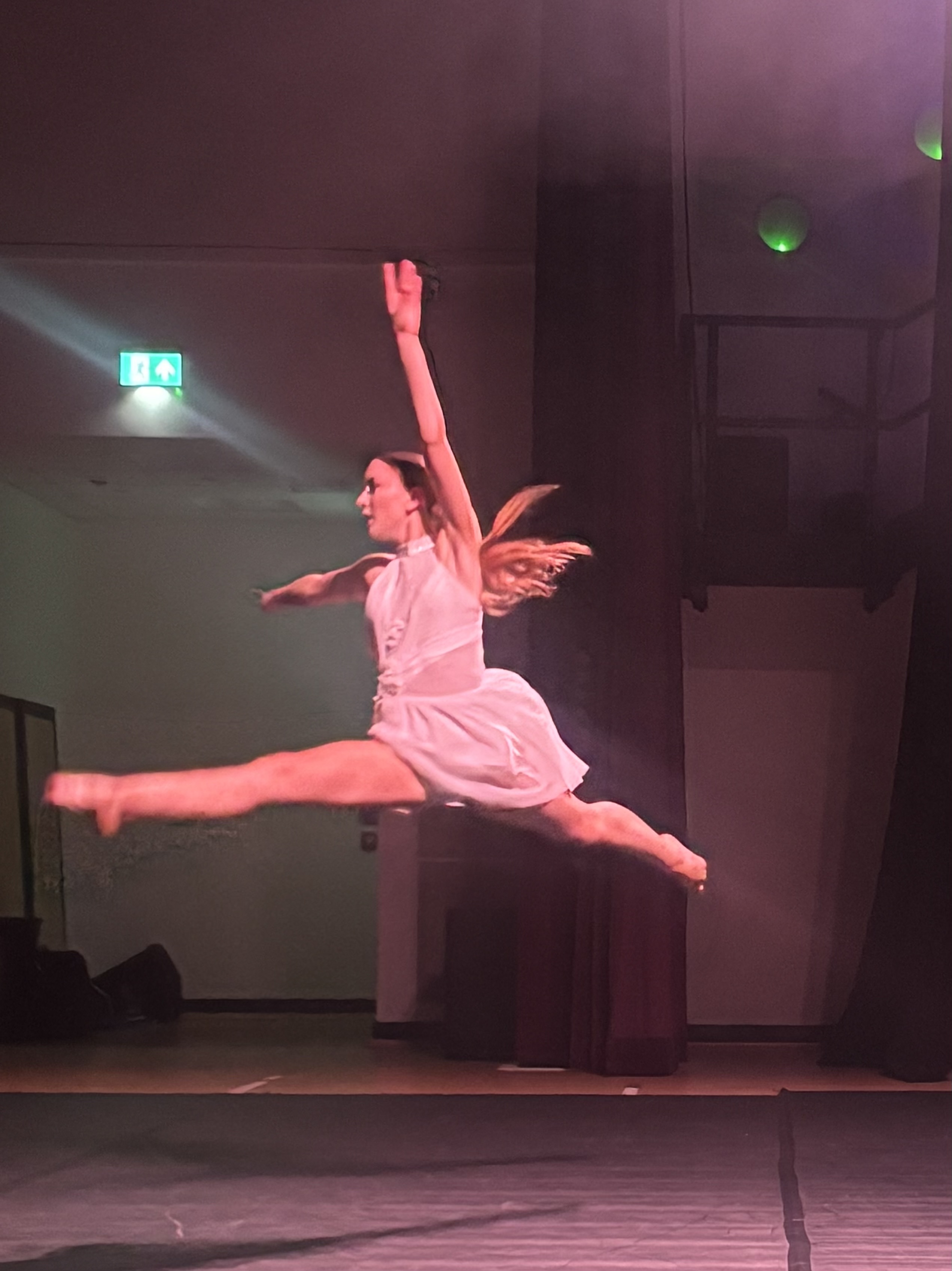 Photograph of a dancer performing a grand jeté leap on stage, wearing a white dress with arms and legs fully extended. The background includes stage curtains, dim lighting, and an illuminated exit sign, highlighting a theatrical performance setting.