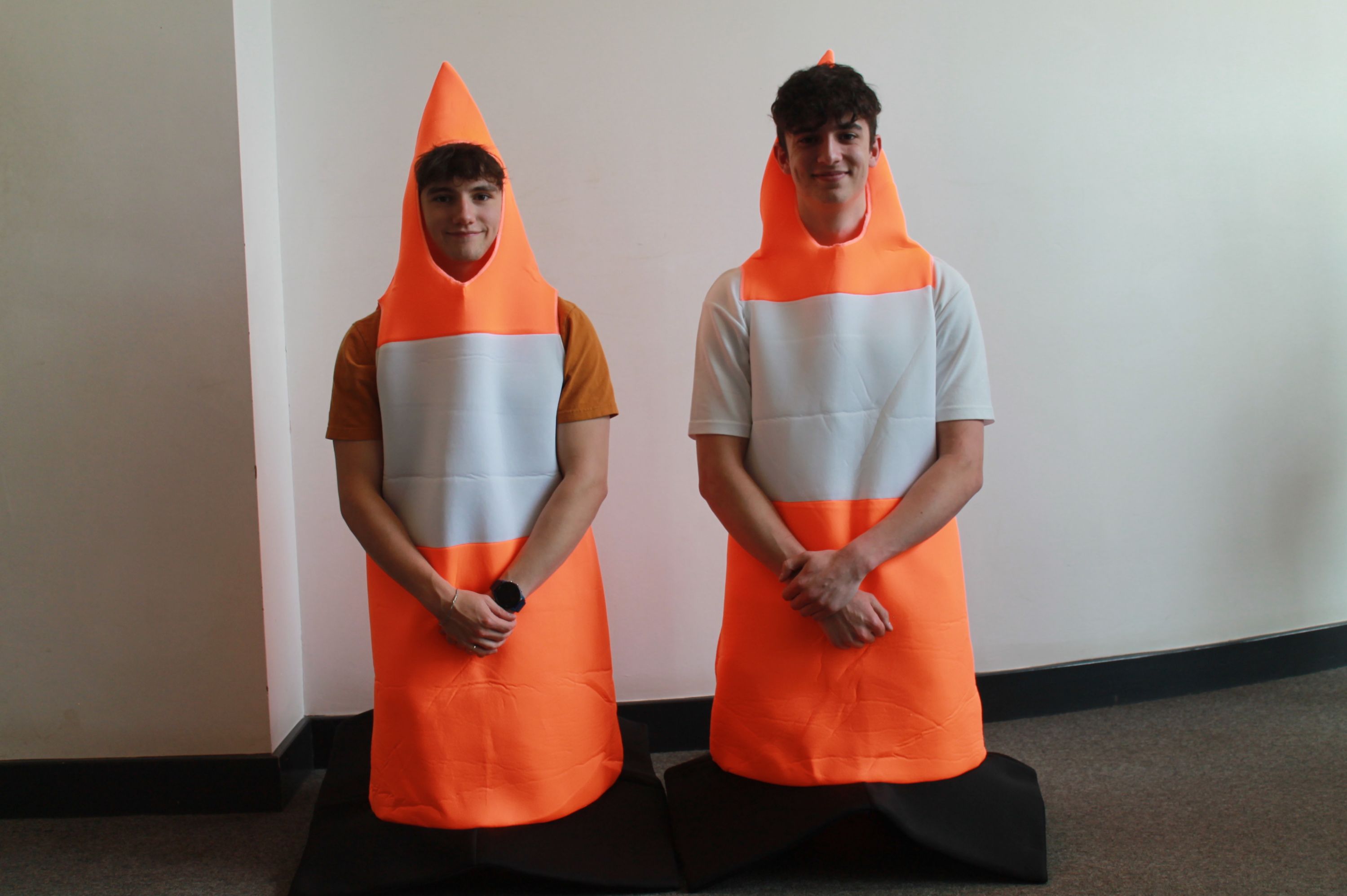 Photograph of two people dressed in bright orange traffic cone costumes standing indoors against a plain white wall. Costumes feature black bases and white reflective stripes, with individuals posing side by side .