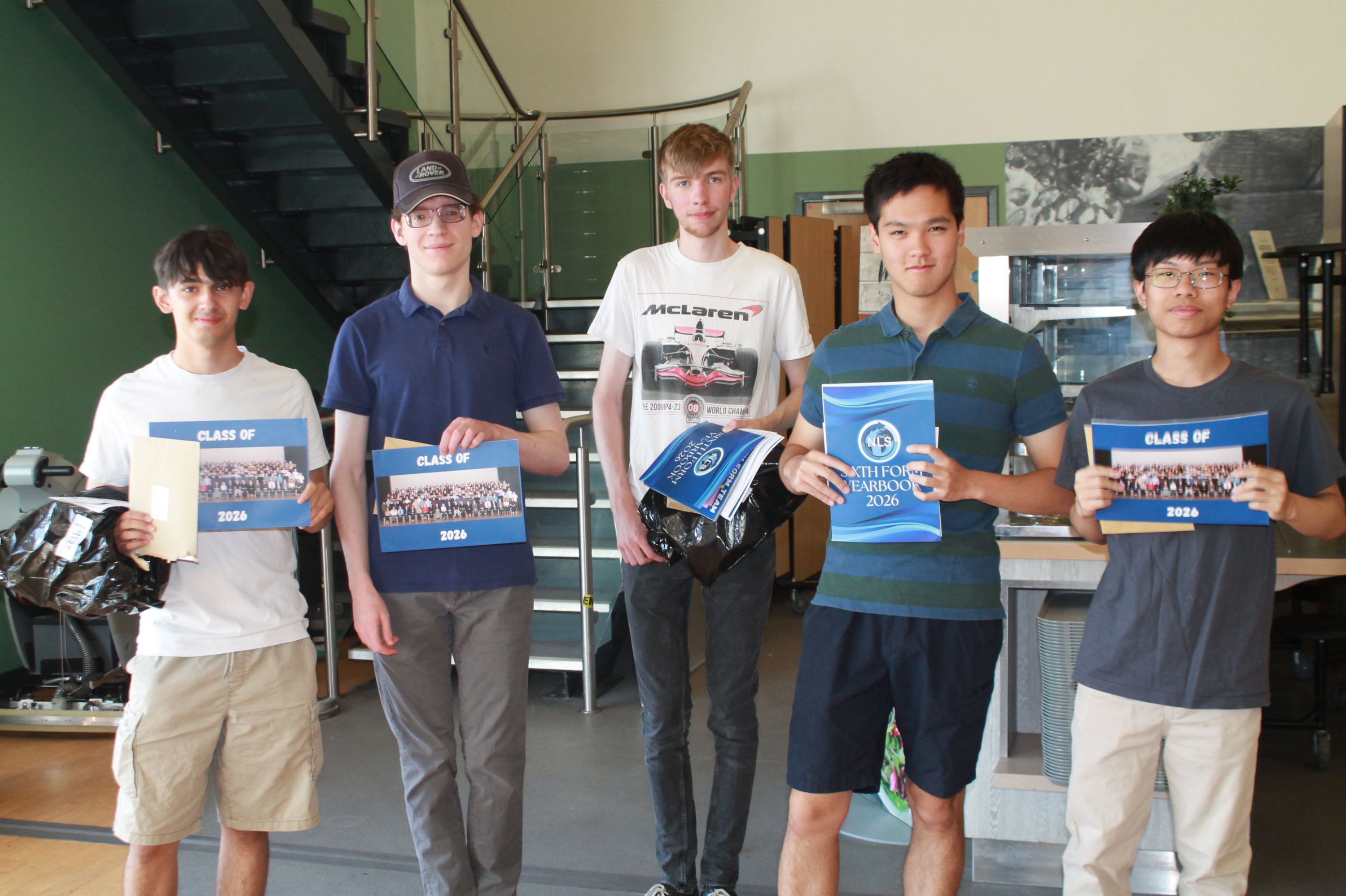 Photograph of five young men standing indoors holding certificates and yearbooks "Class of 2026" , having received their A Level results, background includes a staircase, railing, and modern interior elements