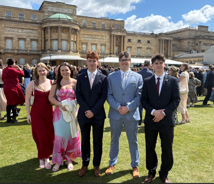 Photograph of a formal outdoor gathering on a lawn in front of a large historic building under a partly cloudy sky. Five young adults dressed in formal attire stand in the foreground, with a crowd of similarly dressed people mingling in the background.