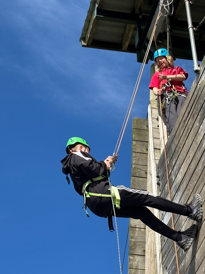 Photograph showing two people engaged in a climbing or abseiling activity on a wooden structure against a clear blue sky. One person is suspended mid-air wearing a green helmet and black clothing, while the other person, wearing a blue helmet and red top, is standing on the platform managing the ropes.
