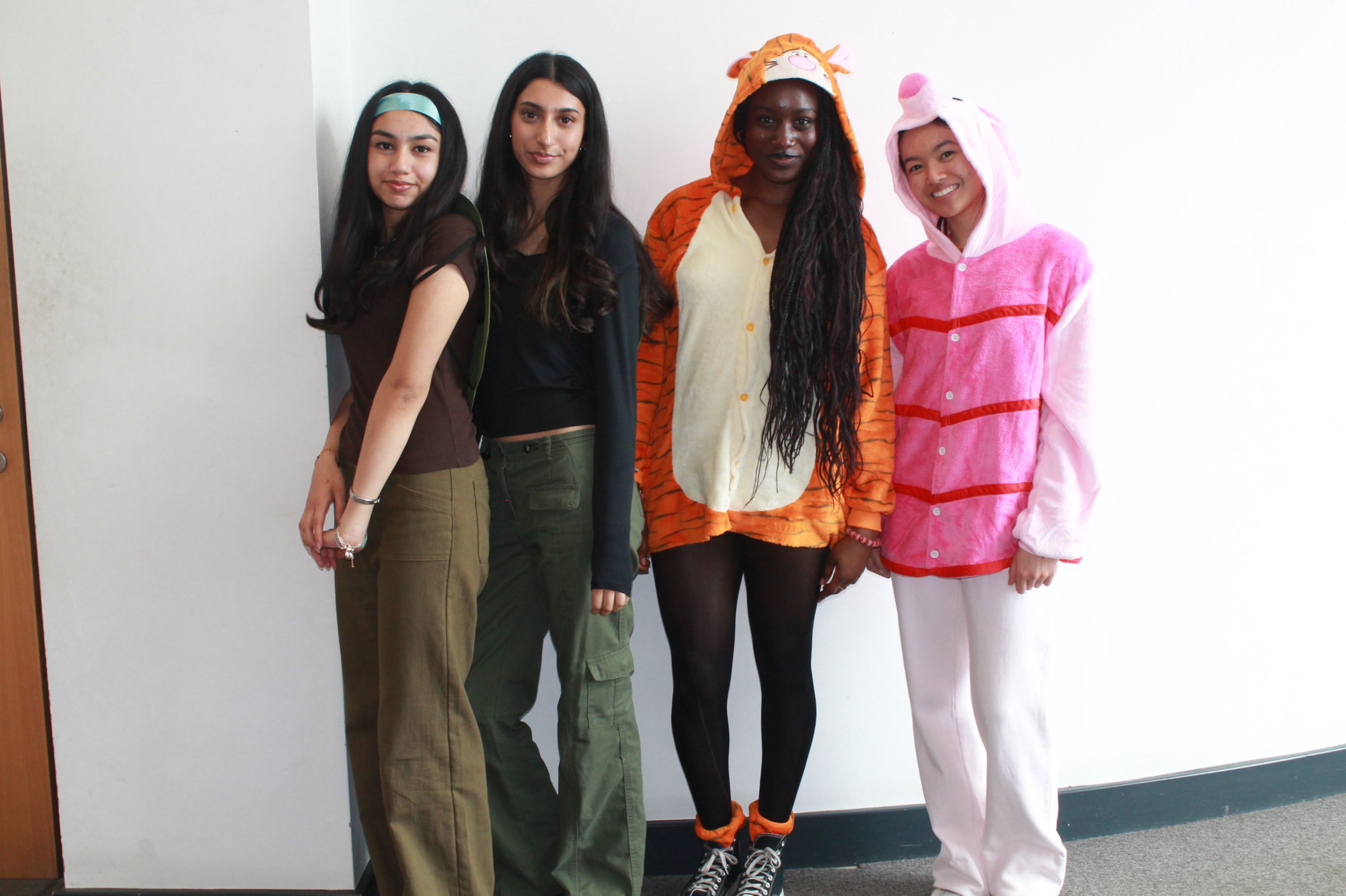 Photograph showing four people standing indoors against a white wall, with two dressed in casual clothing and two wearing animal-themed onesies in orange and pink.