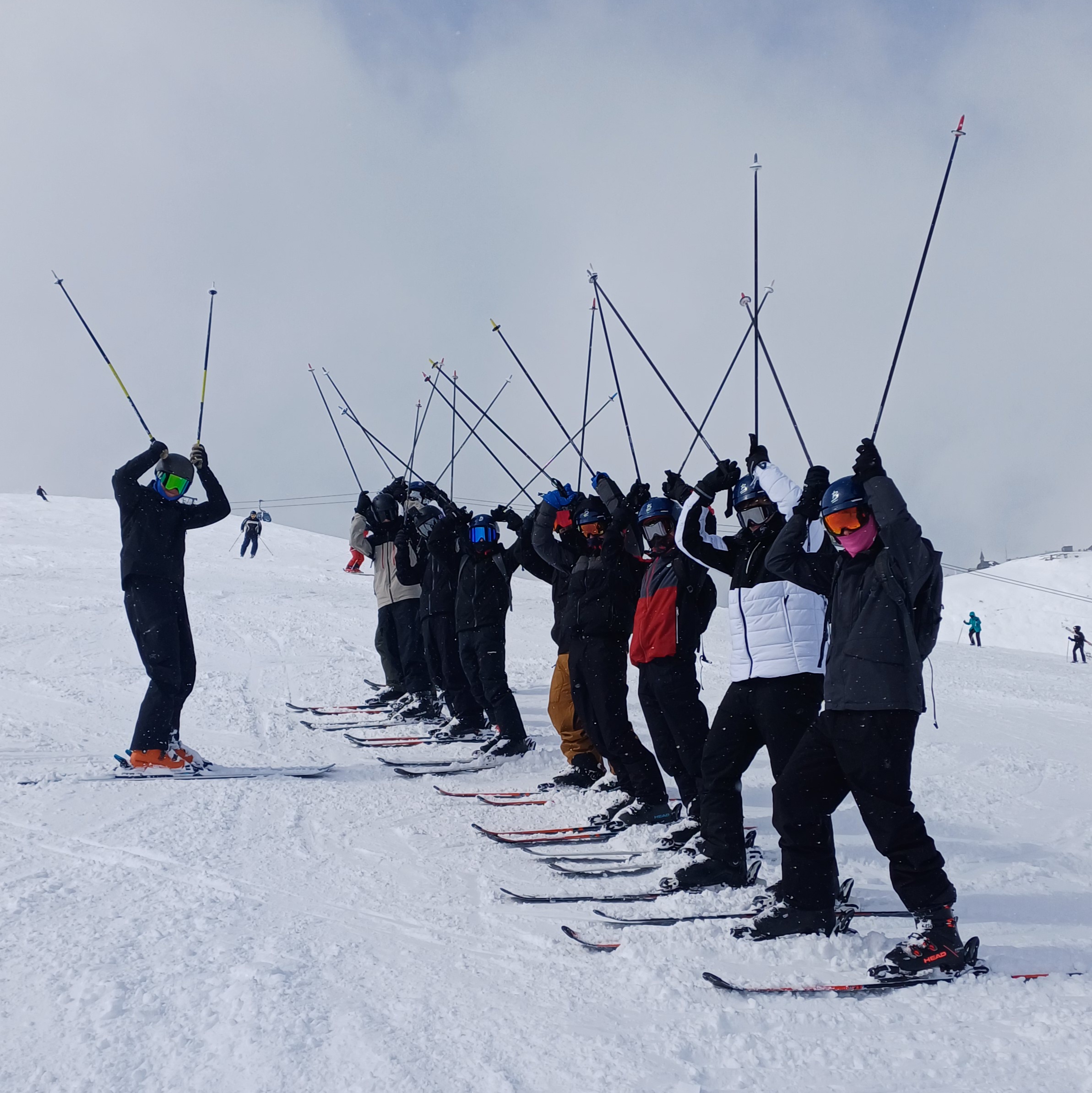 Photograph of a group ski lesson on a snowy slope with an instructor leading several skiers standing in a line, all raising ski poles above heads. Participants wear various coloured ski jackets and helmets, with snowy mountain and cloudy sky background, indicating a training or warm-up session.