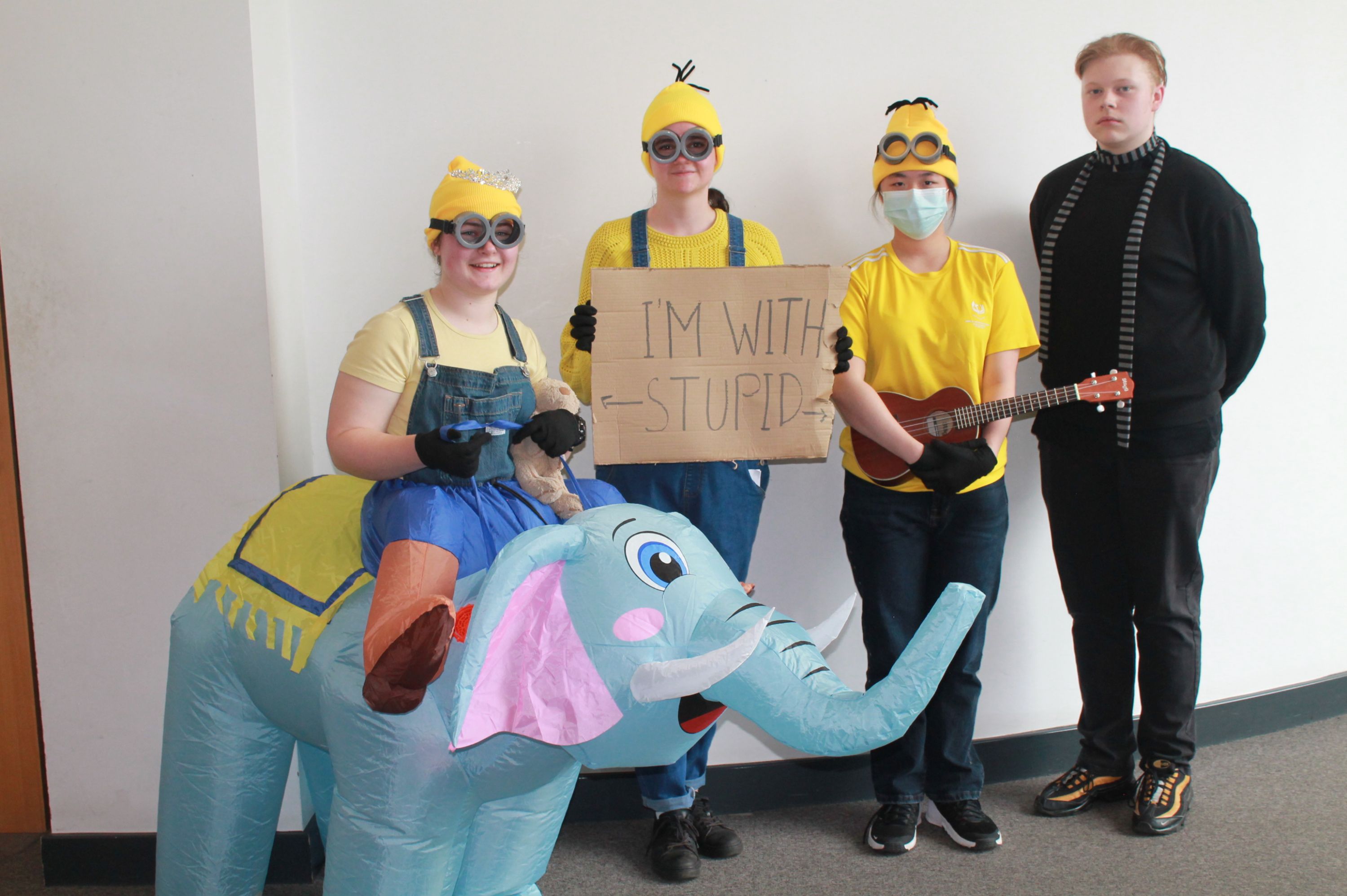 Four people stand against a plain wall, three dressed in yellow and blue costumes resembling Minions, with one holding a cardboard sign reading "I'M WITH STUPID." One person wears an inflatable elephant costume, another holds a small guitar, and the fourth person is dressed in black with a striped scarf.