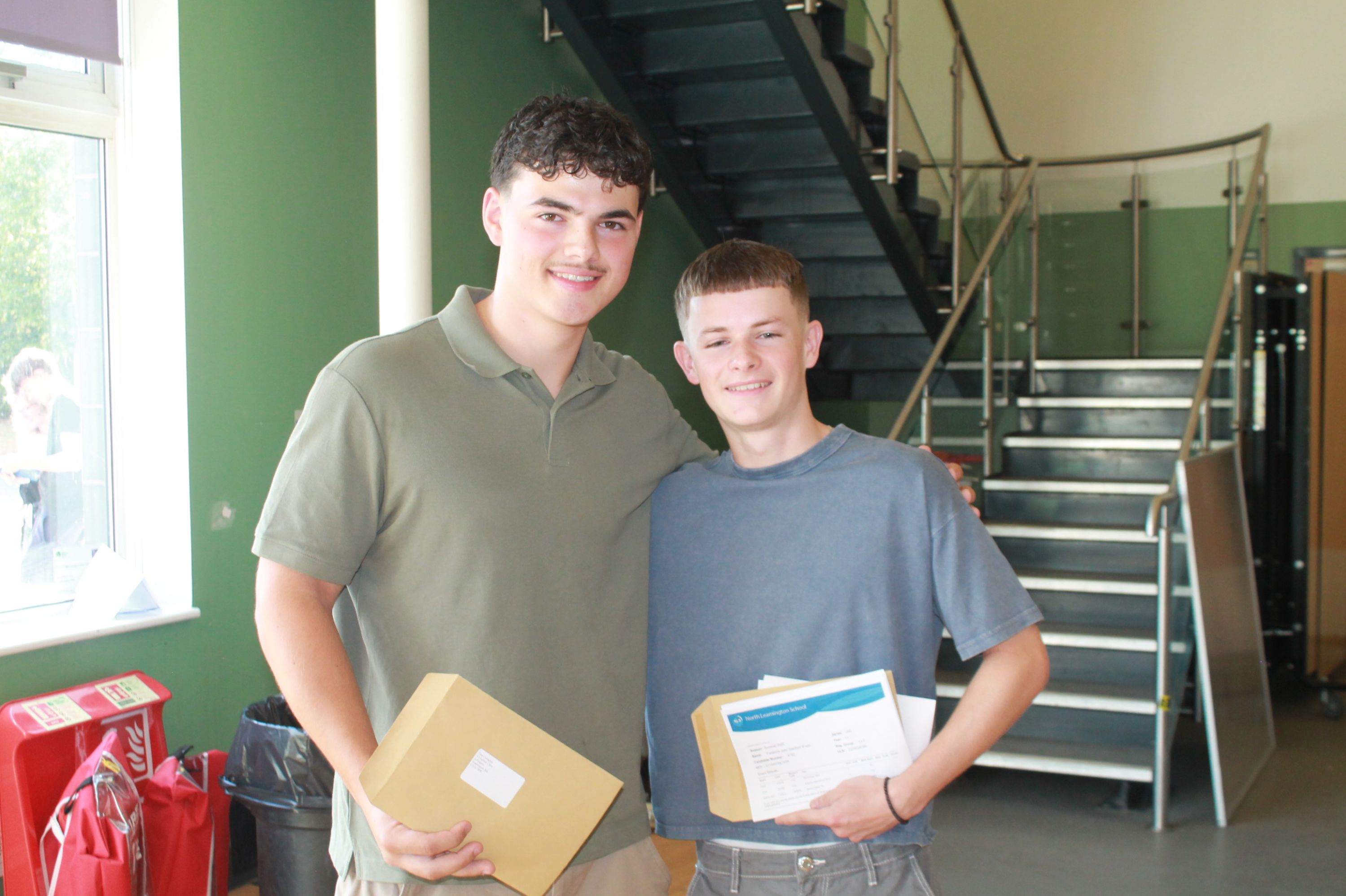 ​​Photograph showing two young women smiling and holding A level results standing indoors near a staircase with green walls in background