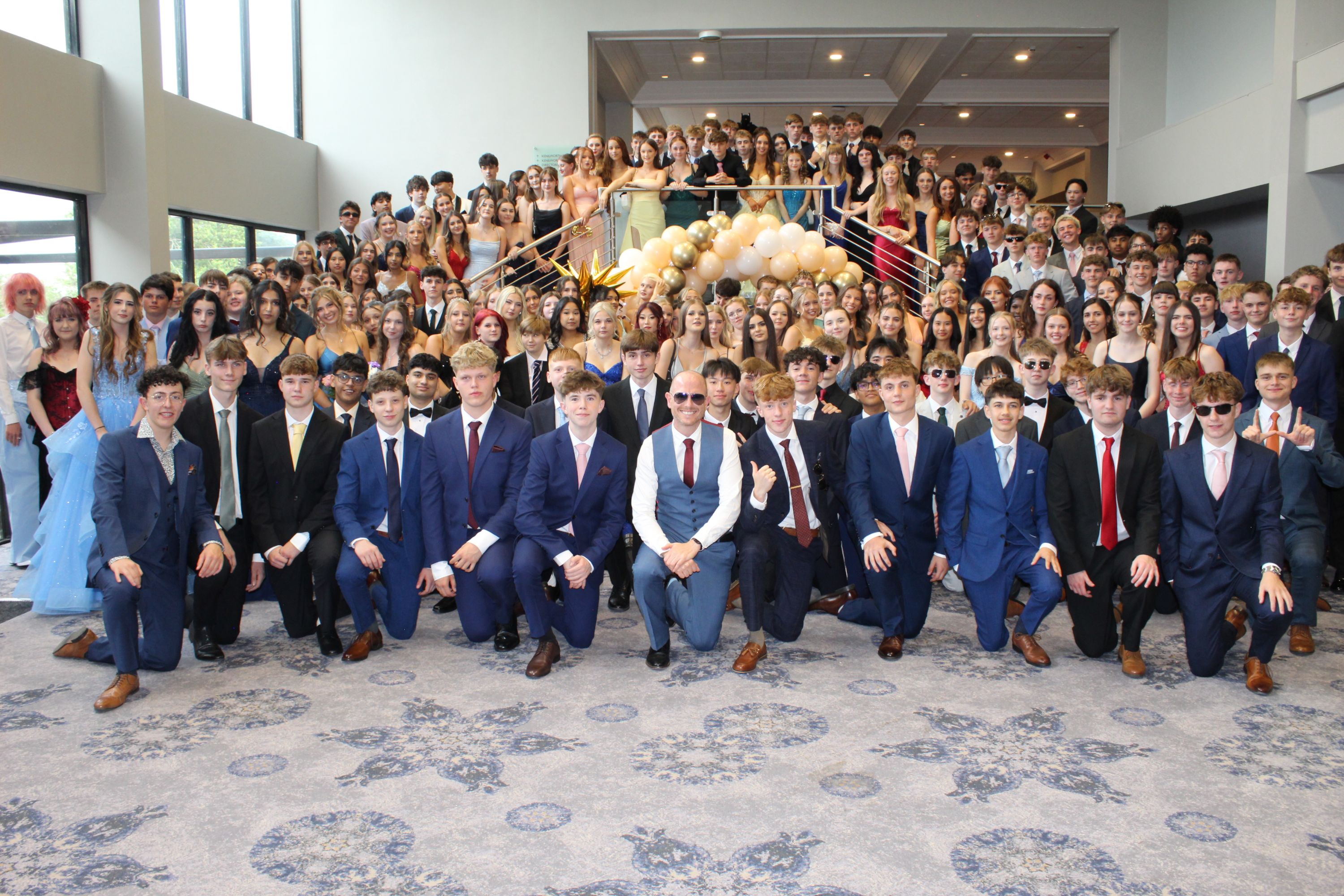 Photograph of a large group of young people dressed formally, gathered indoors on a carpeted floor and staircase decorated with gold and white balloons. Group includes individuals in suits, dresses, and sunglasses, at prom .