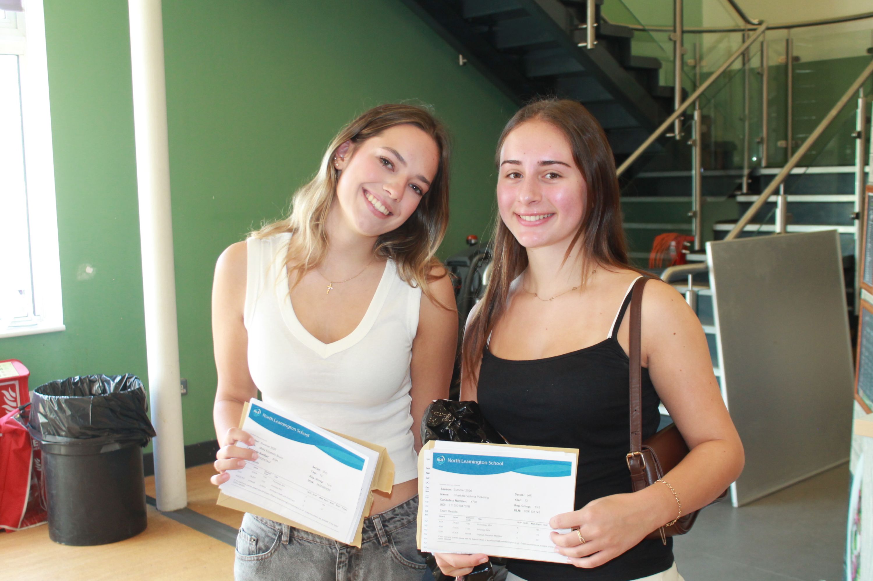 ​​Photograph showing two young women smiling and holding A level results standing indoors near a staircase with green walls in background. Both wear casual tops, one white and one black, 