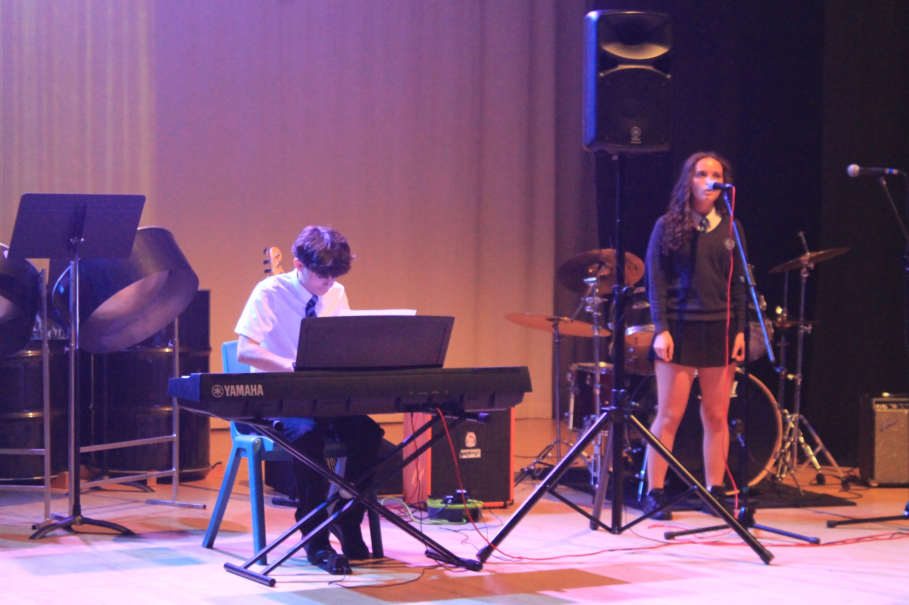 Photograph of a musical performance featuring a pianist playing a Yamaha keyboard and a female vocalist singing into a microphone on stage. Stage setup includes a drum kit, music stands, and speakers, with warm lighting highlighting performers against a plain backdrop.