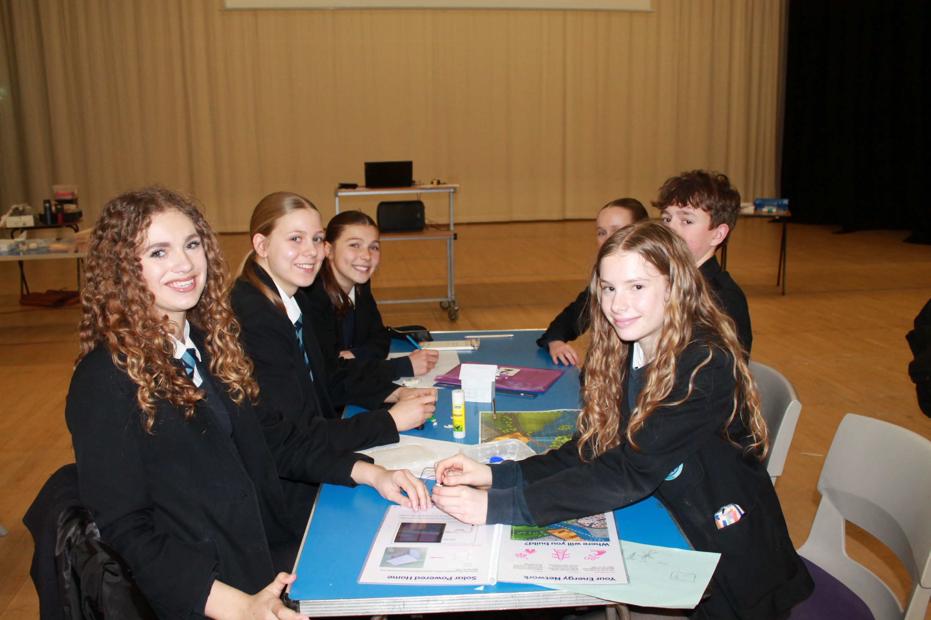 Photograph of six students in school uniforms seated around a blue table engaged in a group activity involving papers and glue sticks. Setting is a  hall with beige curtains and additional tables in the background