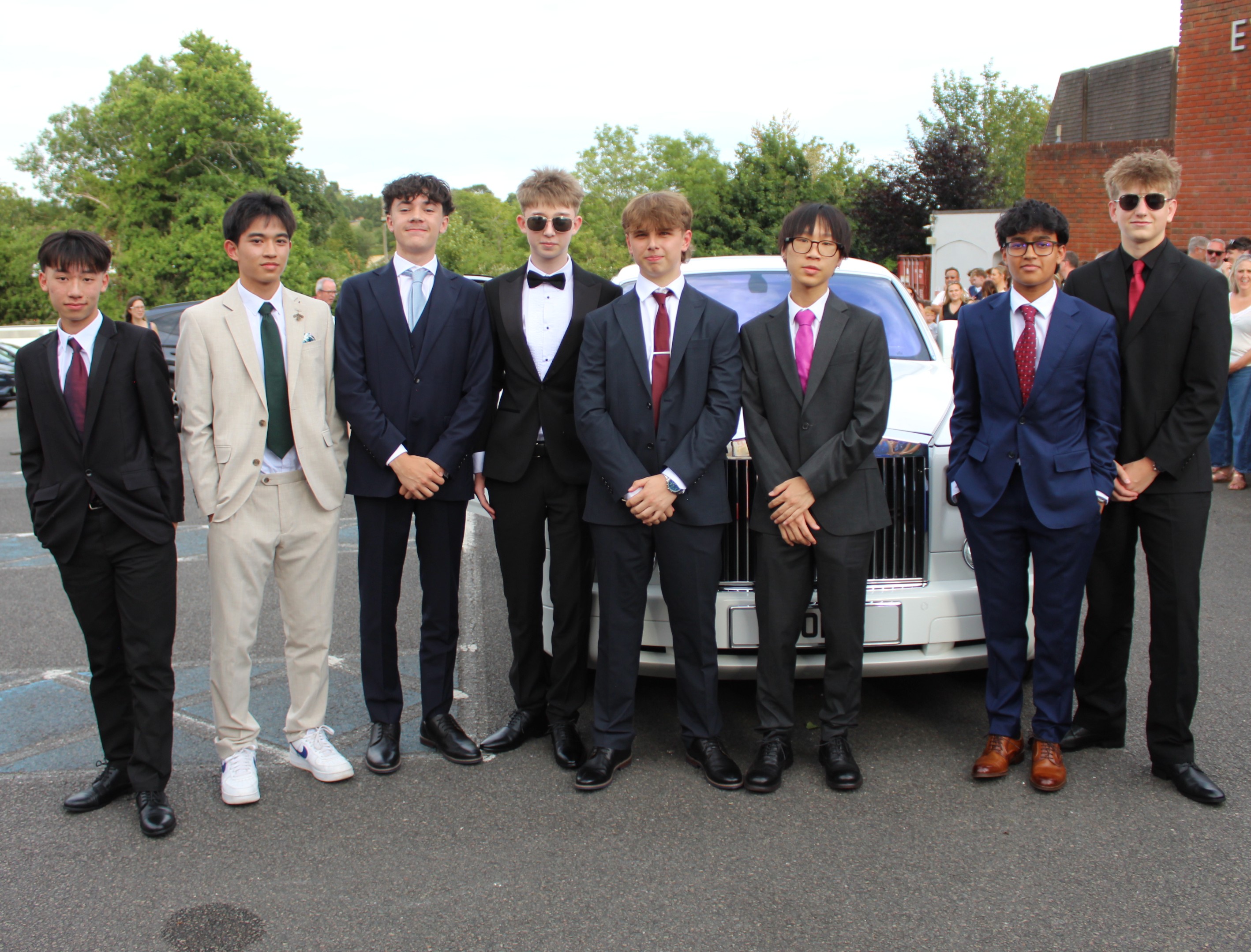Photograph of eight young men dressed in formal suits standing in front of a white luxury car outdoors. The group displays a variety of suit styles and colours, with some wearing ties and others bow ties, at prom.