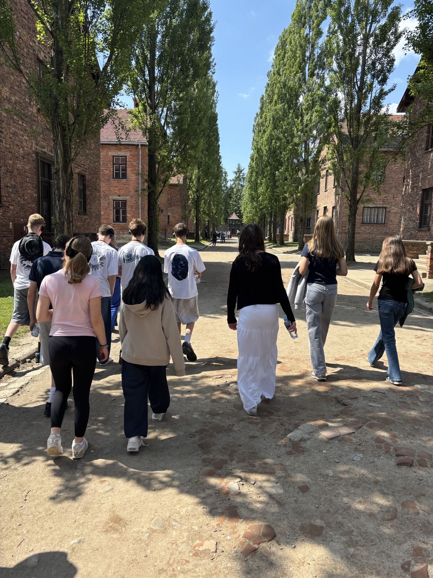 Photograph showing a group of people walking along a sunlit path between tall brick buildings and trees. The scene highlights casual clothing styles and shadows cast on the ground, suggesting a relaxed outdoor setting on a clear day.