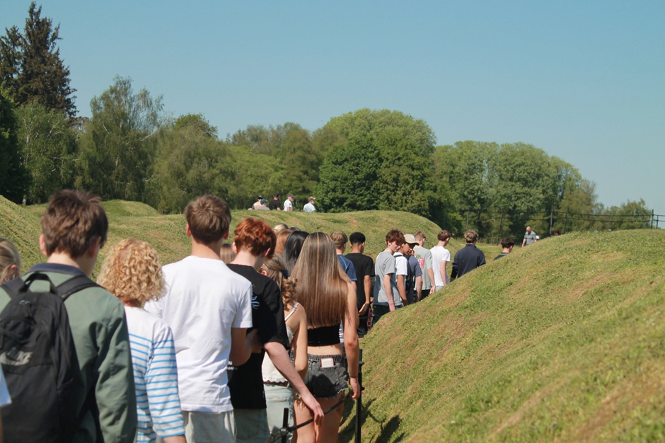 Photograph showing a group of people walking in a line in trenches at Beaumont Hamel, a grassy, undulating path bordered by low hills under a clear blue sky. 
