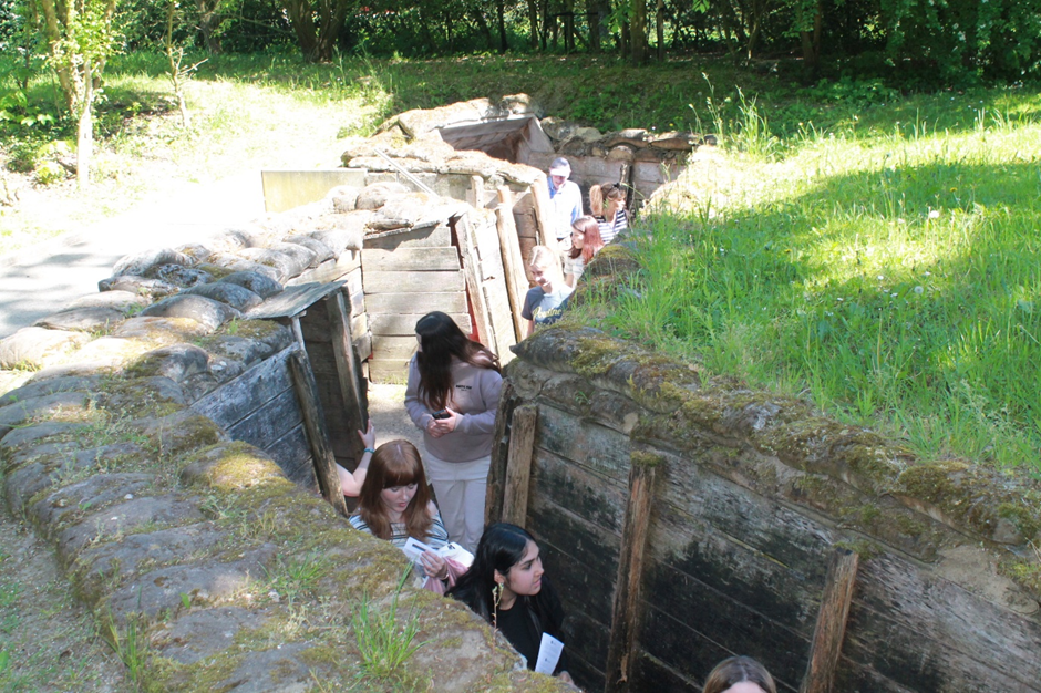 Photograph showing a group of people walking through trenches at the Passchendaele Museum - a narrow, grass-covered trench with wooden reinforcements, part of a historical or war memorial site. Sunlight casts shadows on the trench walls, highlighting the contrast between natural surroundings and man-made structure.