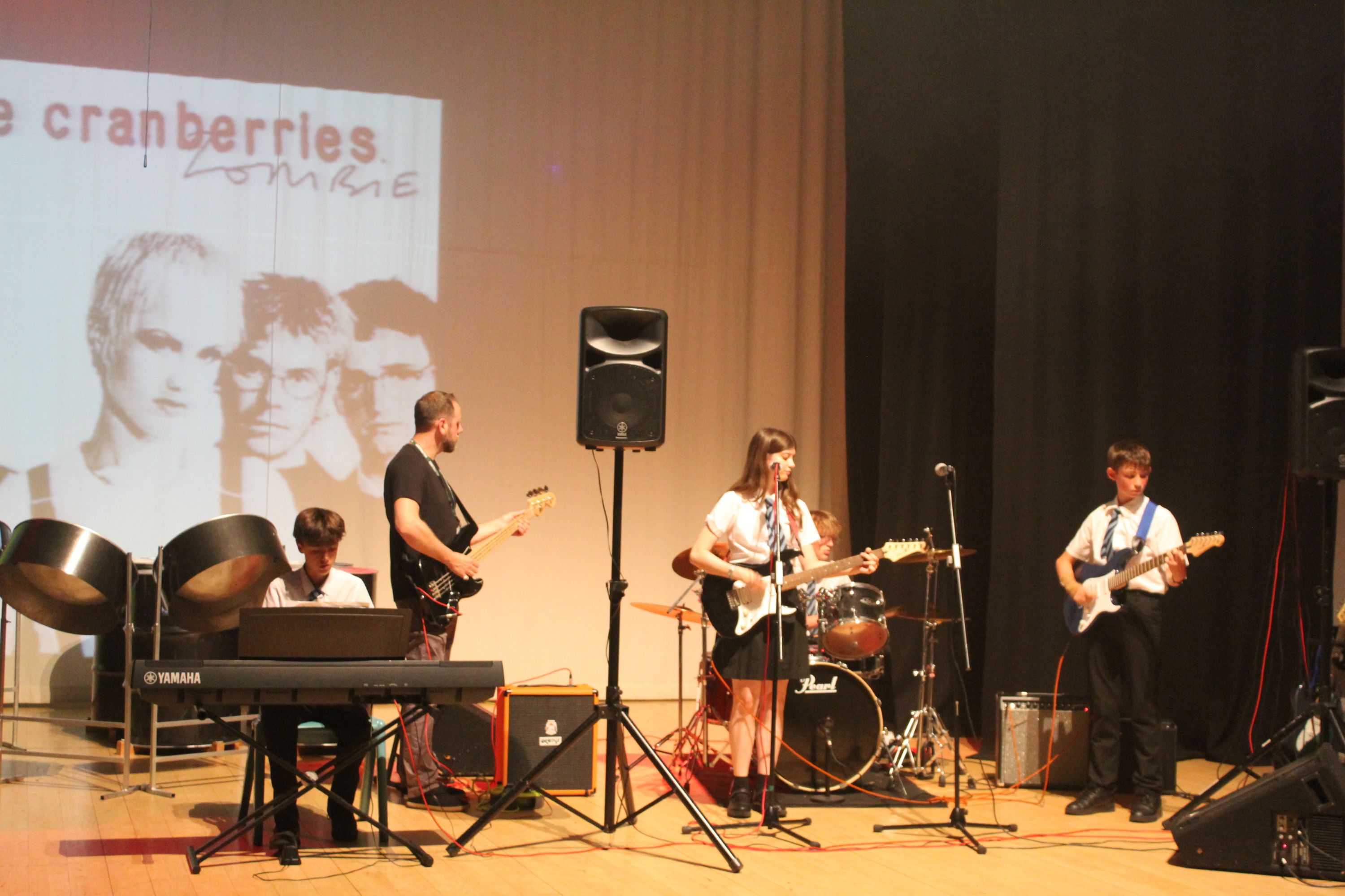 Photograph of a school band performing on stage with instruments including keyboard, electric guitars, and drums, with a projection of The Cranberries album cover in the background. The scene highlights a live music event, featuring four young musicians dressed in school uniforms, with microphones and amplifiers set up for the performance.