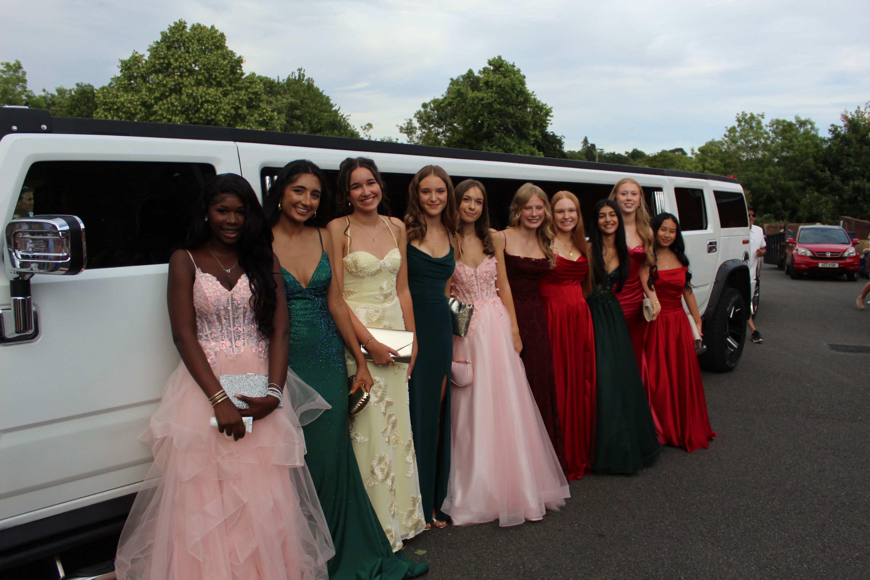 photograph of ten young women dressed in formal evening gowns standing in a row beside a white stretch limousine, suggesting a special event like prom or a formal celebration. Dresses feature various colours including pink, green, cream, and red, with greenery and parked cars visible in background under a cloudy sky.