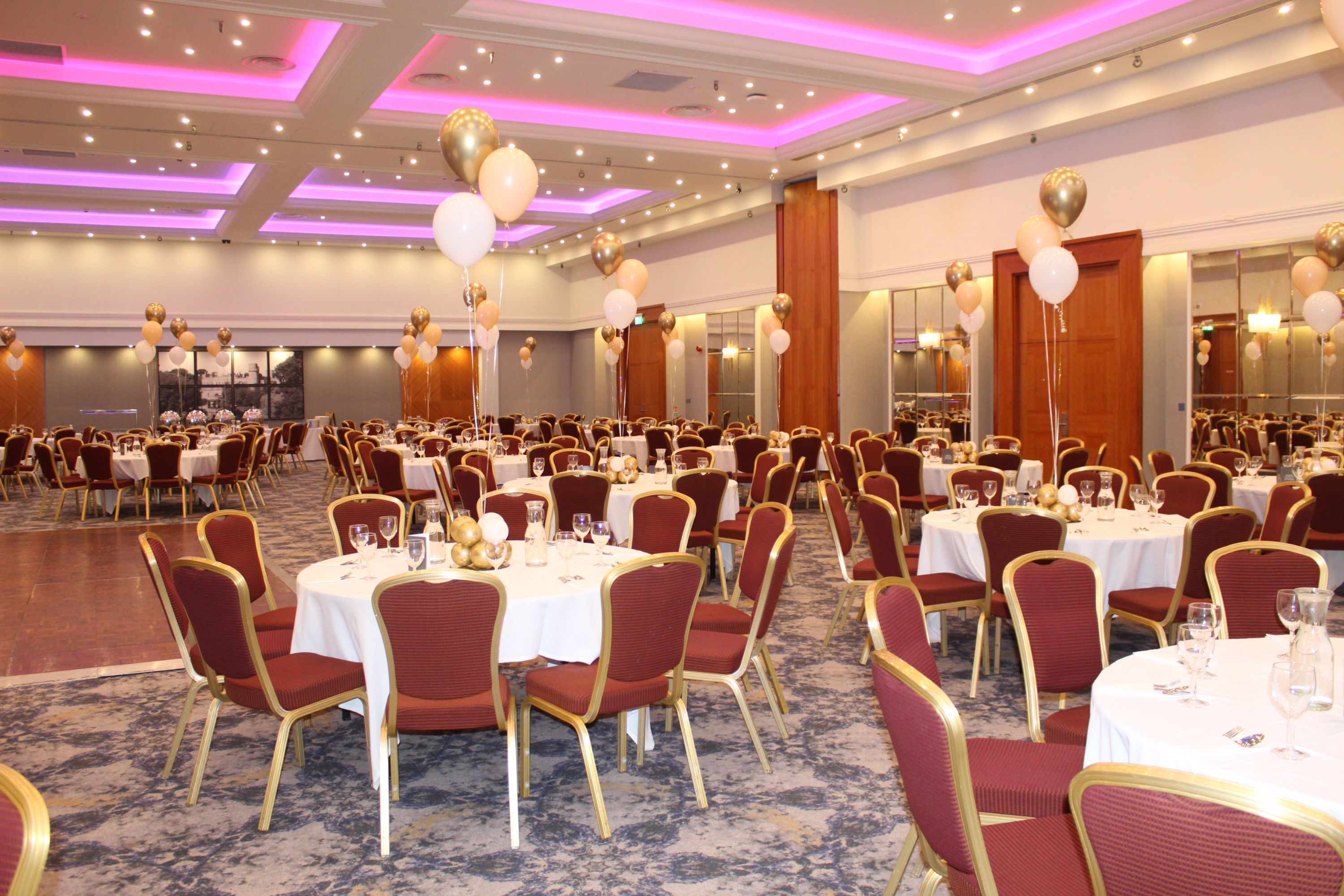 Photograph of a large banquet hall set up for prom with round tables covered in white tablecloths and surrounded by red cushioned chairs. Gold and white balloons decorate tables, while soft purple lighting illuminates ceiling panels, creating an elegant atmosphere.