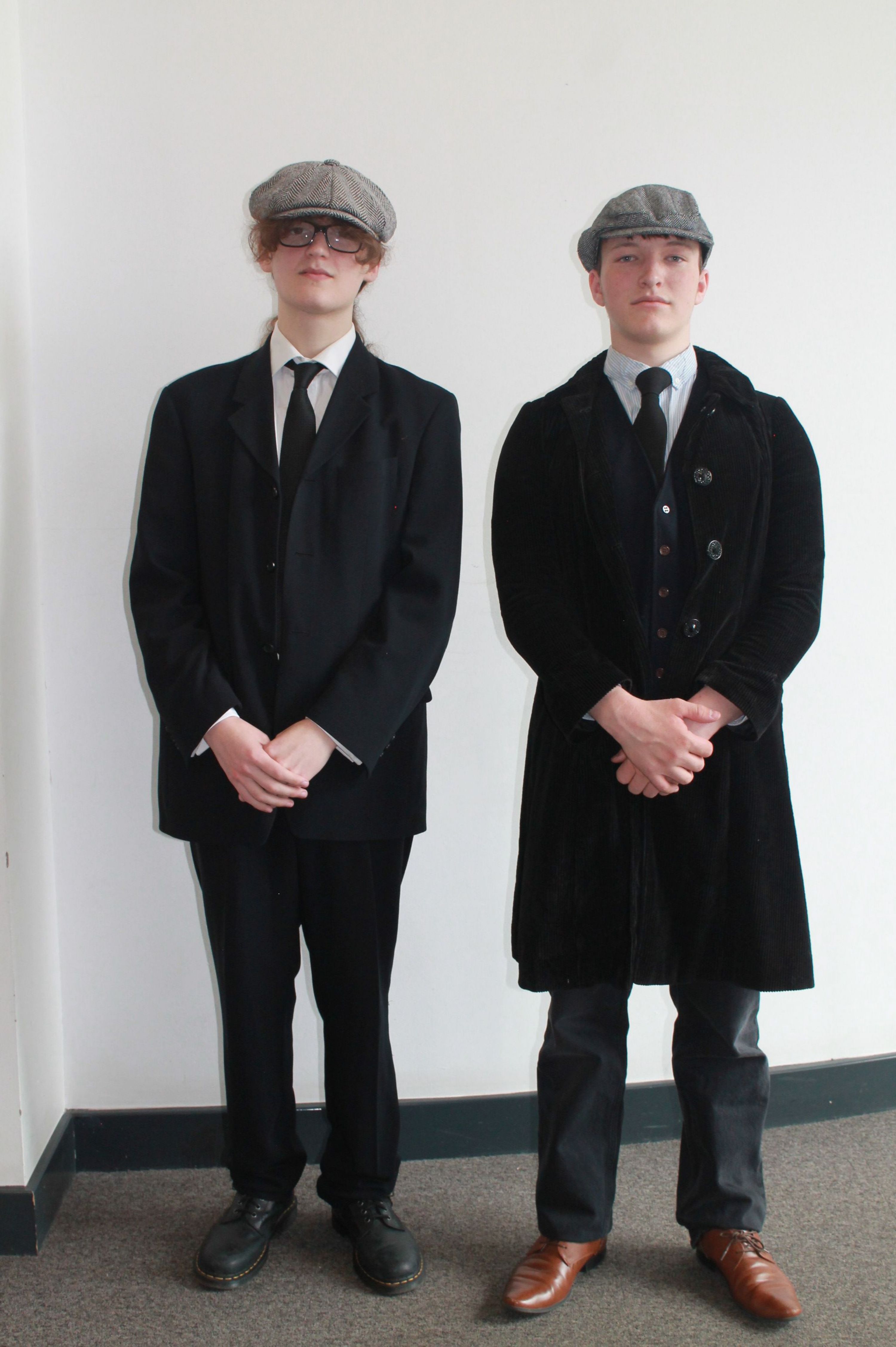 Photograph of two people standing side by side against plain white wall, both wearing black suits, white shirts, black ties, and grey flat caps. One wears brown shoes and a black velvet coat, while the other wears black shoes and a black blazer.