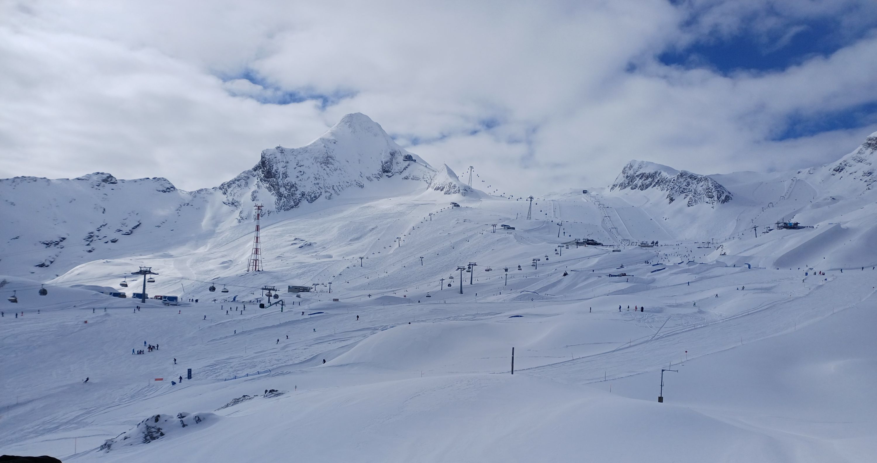 Photograph of a snow-covered mountain ski resort under a partly cloudy sky, featuring ski lifts and scattered skiers on various slopes. Prominent peaks and ridges form a dramatic backdrop, with ski infrastructure including poles and cables visible across the terrain.