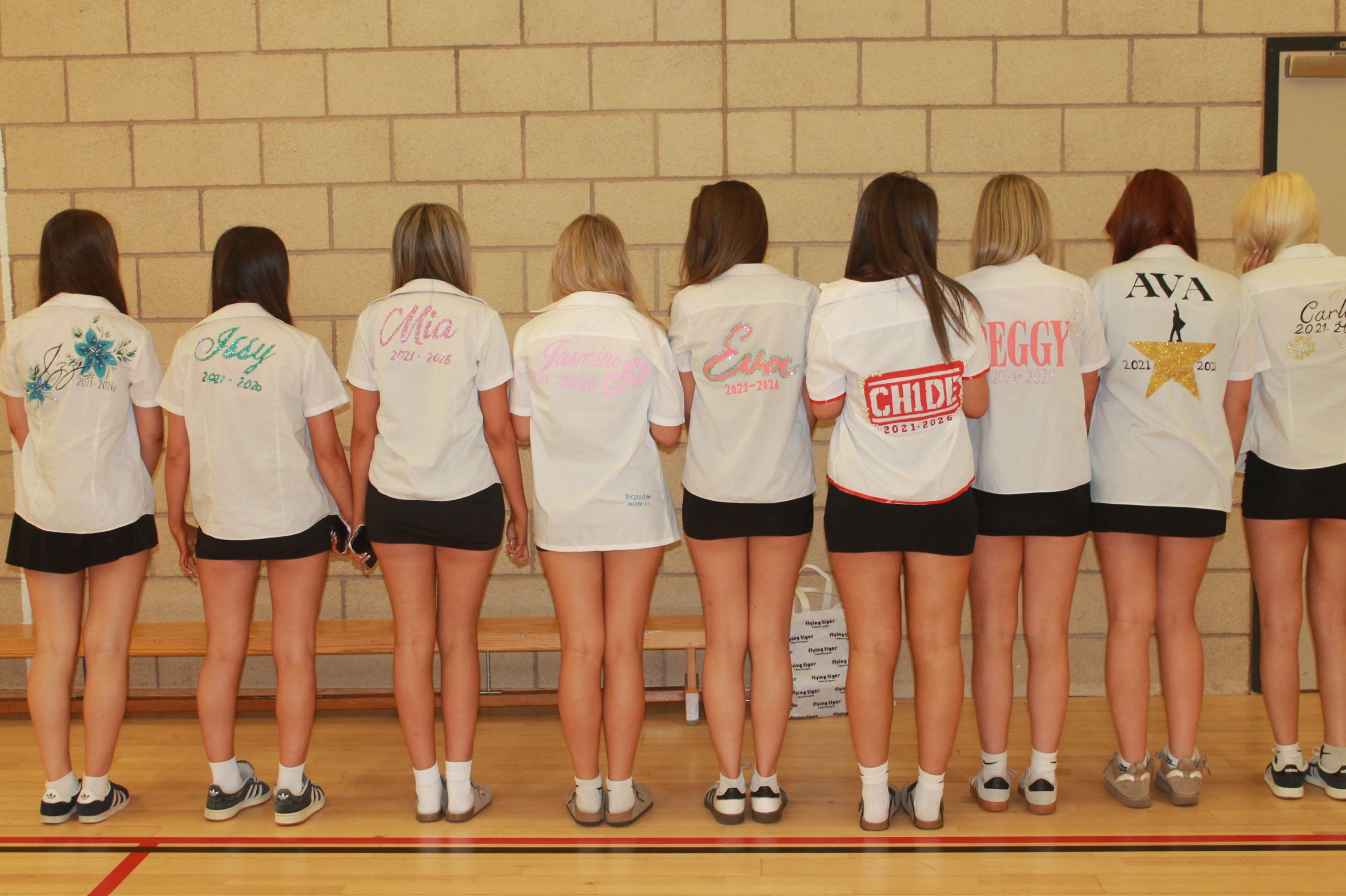 Photograph of nine girls standing in a row facing away, wearing personalised white shirts with names and designs on backs, paired with black shorts or skirts. Shirts feature various colours and fonts, including names like "Chloe," "Ava," and "Ellie," with some shirts displaying additional symbols such as stars and logos.