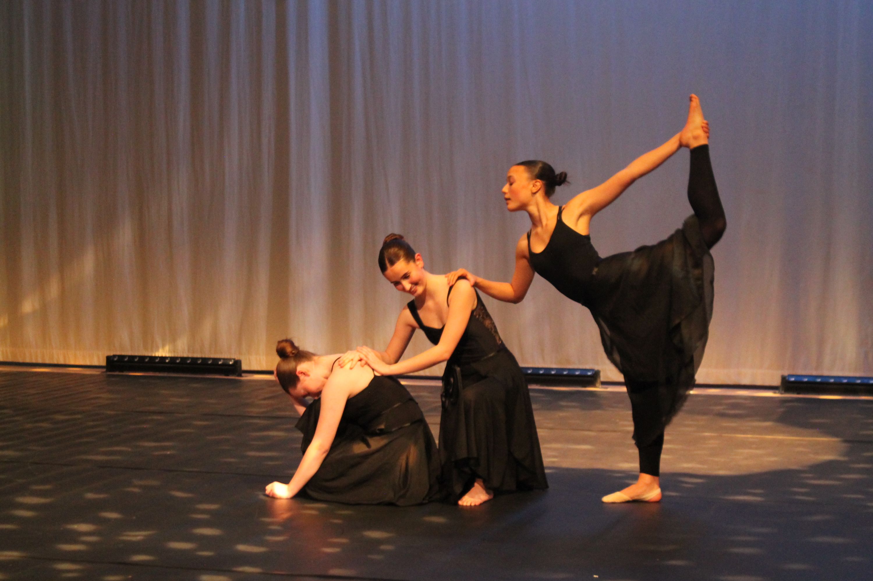 Photograph of three dancers performing on stage wearing black dresses against a plain backdrop with stage lights along the floor. One dancer is kneeling with head down, another is standing and supporting the kneeling dancer, and the third is balancing on one leg while holding the other leg extended behind