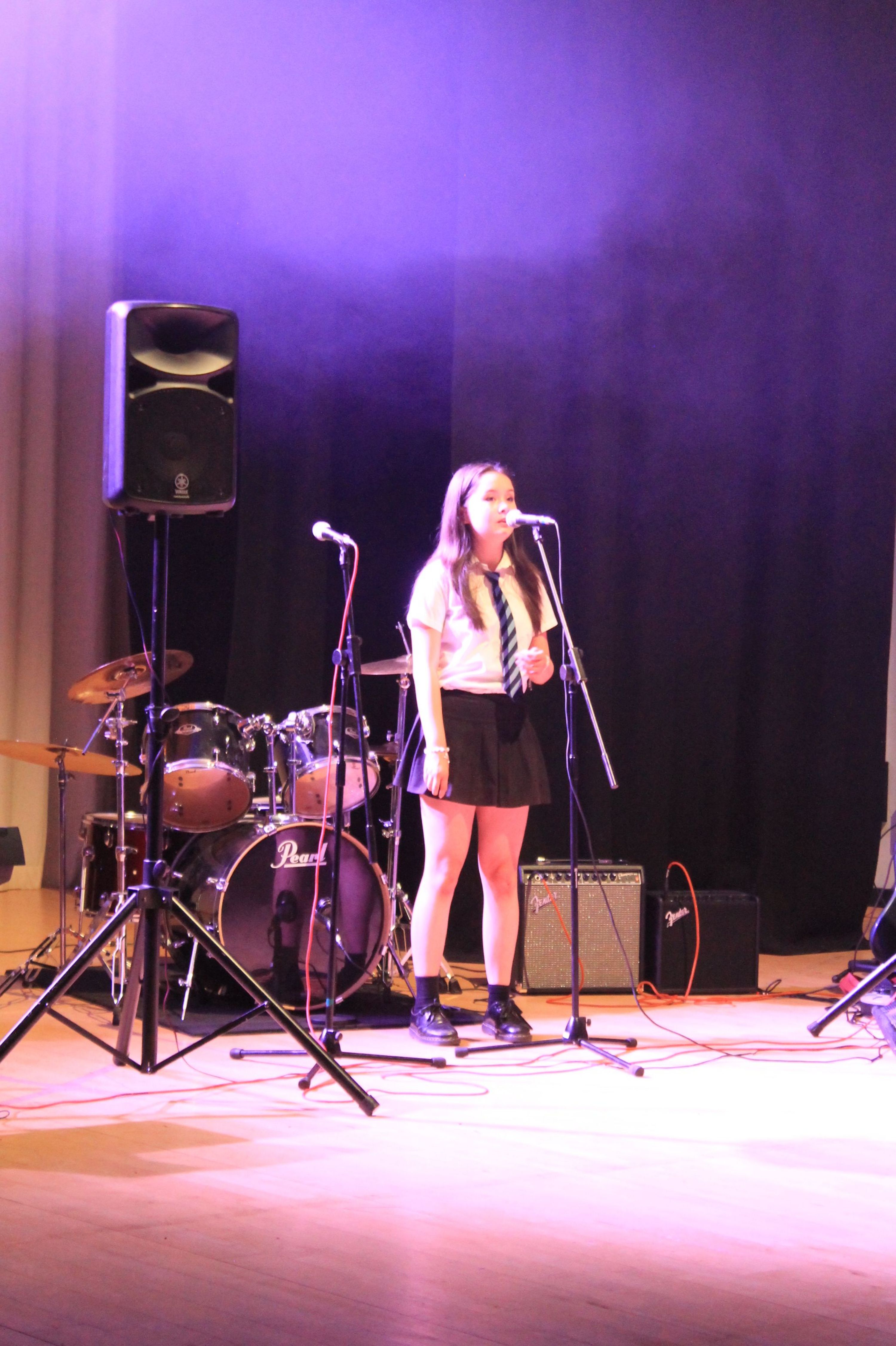 Photograph of a female performer standing on stage with a microphone, surrounded by musical instruments including a drum set and amplifiers. Stage lighting casts a purple hue, highlighting the performer dressed in a dark outfit and white shoes, suggesting a live music or vocal performance event.