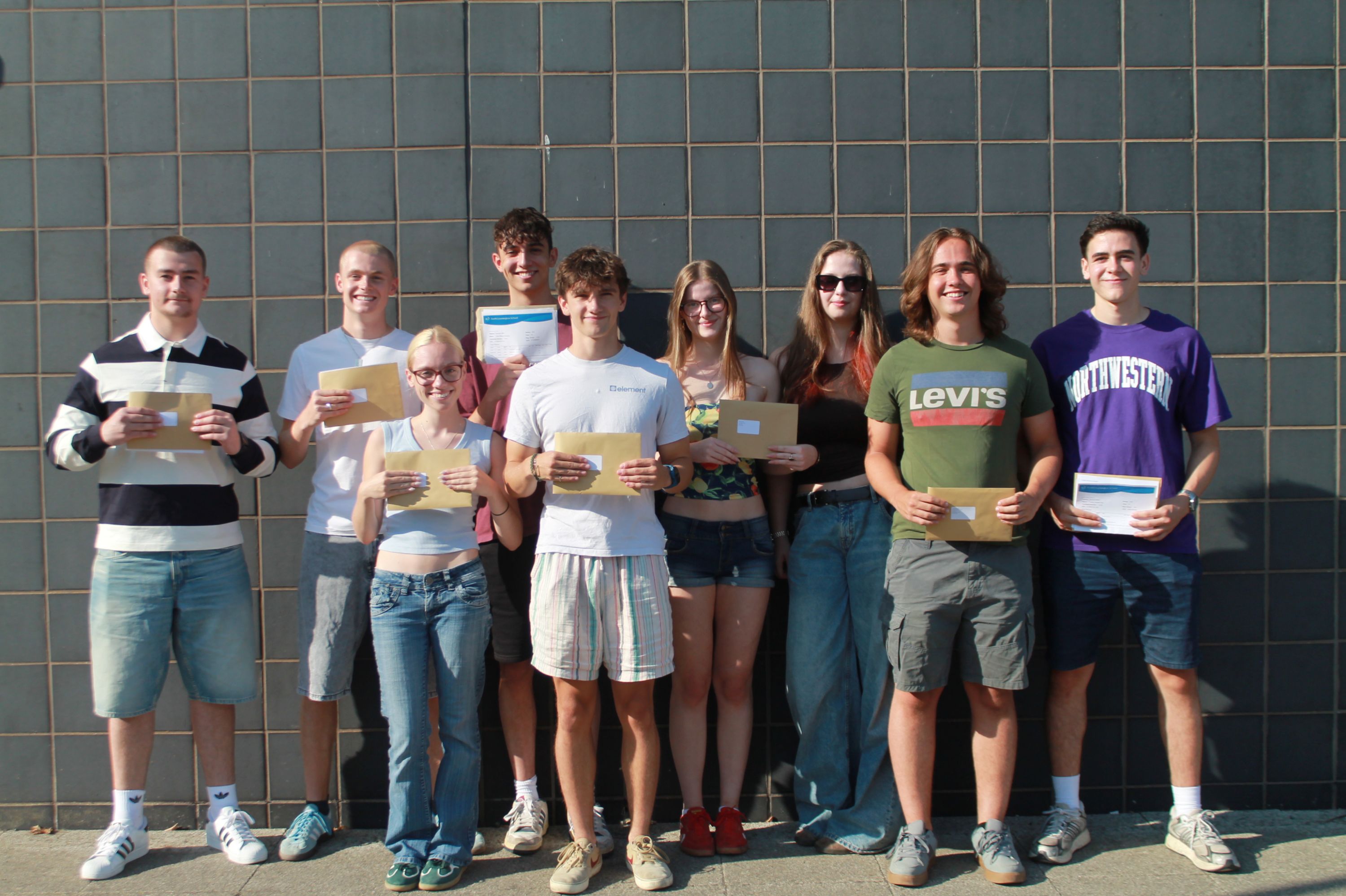   Photograph of nine young people standing in a row against a tiled wall, each holding an envelope or paper of their A level results . The group are celebrating  and  sharing results, with casual clothing and a sunny outdoor setting.