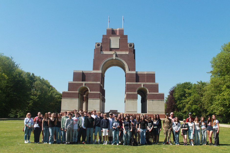 Photograph of a large group of people standing in front of the Thiepval Memorial, (a brick and stone war memorial arch )under a clear blue sky. The memorial features three arches with two flagpoles on top, surrounded by green trees and grass