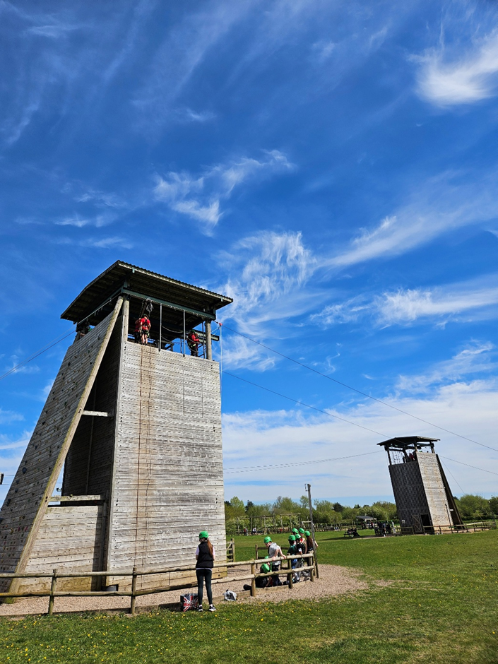 Photograph of an outdoor climbing and zipline structure with people engaging in activities. The tall concrete tower features climbing walls and a zipline platform, set against a bright blue sky with scattered clouds and surrounded by grassy area