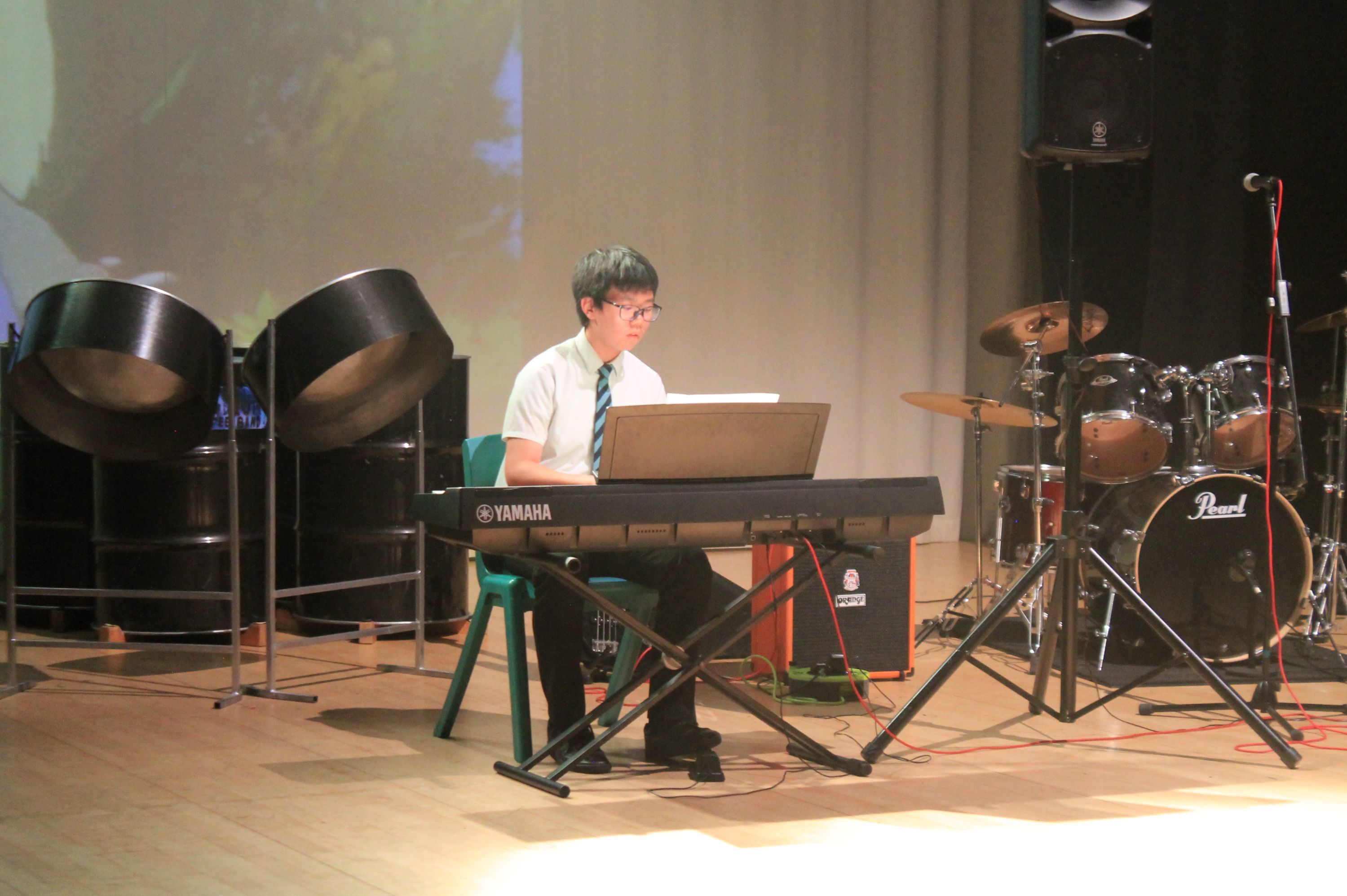 Photograph of a young musician playing a Yamaha keyboard on stage during a performance. Surrounding instruments include steel drums on the left and a Pearl drum set on the right, with stage lighting and sound equipment visible in the background.