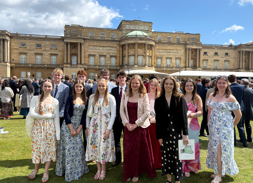 Photograph showing a group of young people dressed formally standing on grass in front of a large historic building under a partly cloudy sky at Buckingham Palace in a formal event or gathering, with individuals wearing suits and dresses in various colours and patterns, and a crowd visible in the background.