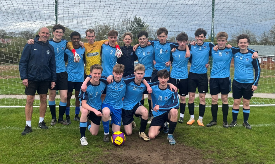 Photograph of a football team posing in front of a goal on a grass field, wearing blue jerseys, black shorts, and black socks. The group includes 15 players and one coach, with one of the players kneeling behind a yellow and purple football, under an overcast sky.